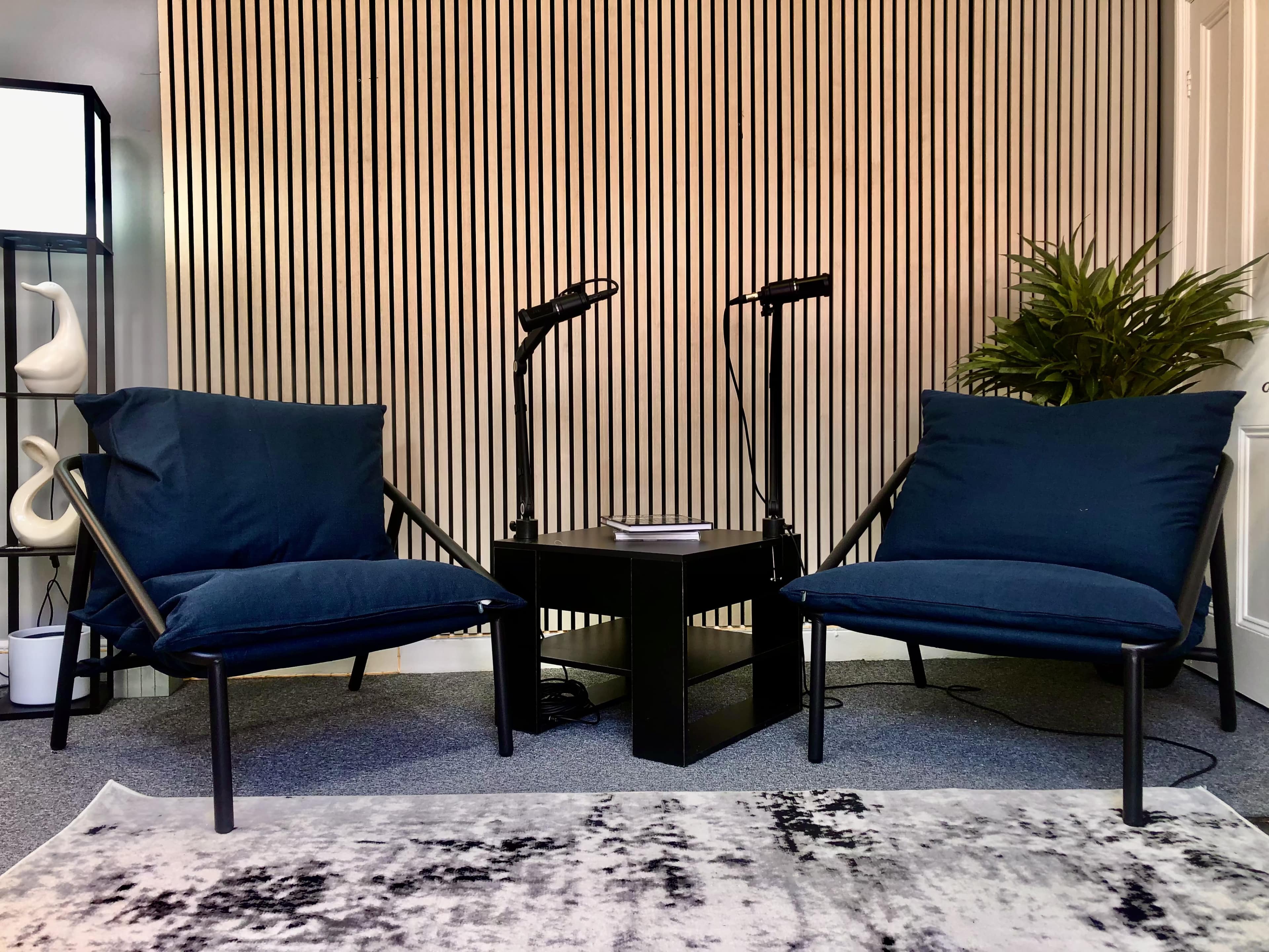 Two navy blue chairs facing each other around a black table, with a potted plant and lamps in the background against a striped wall.
