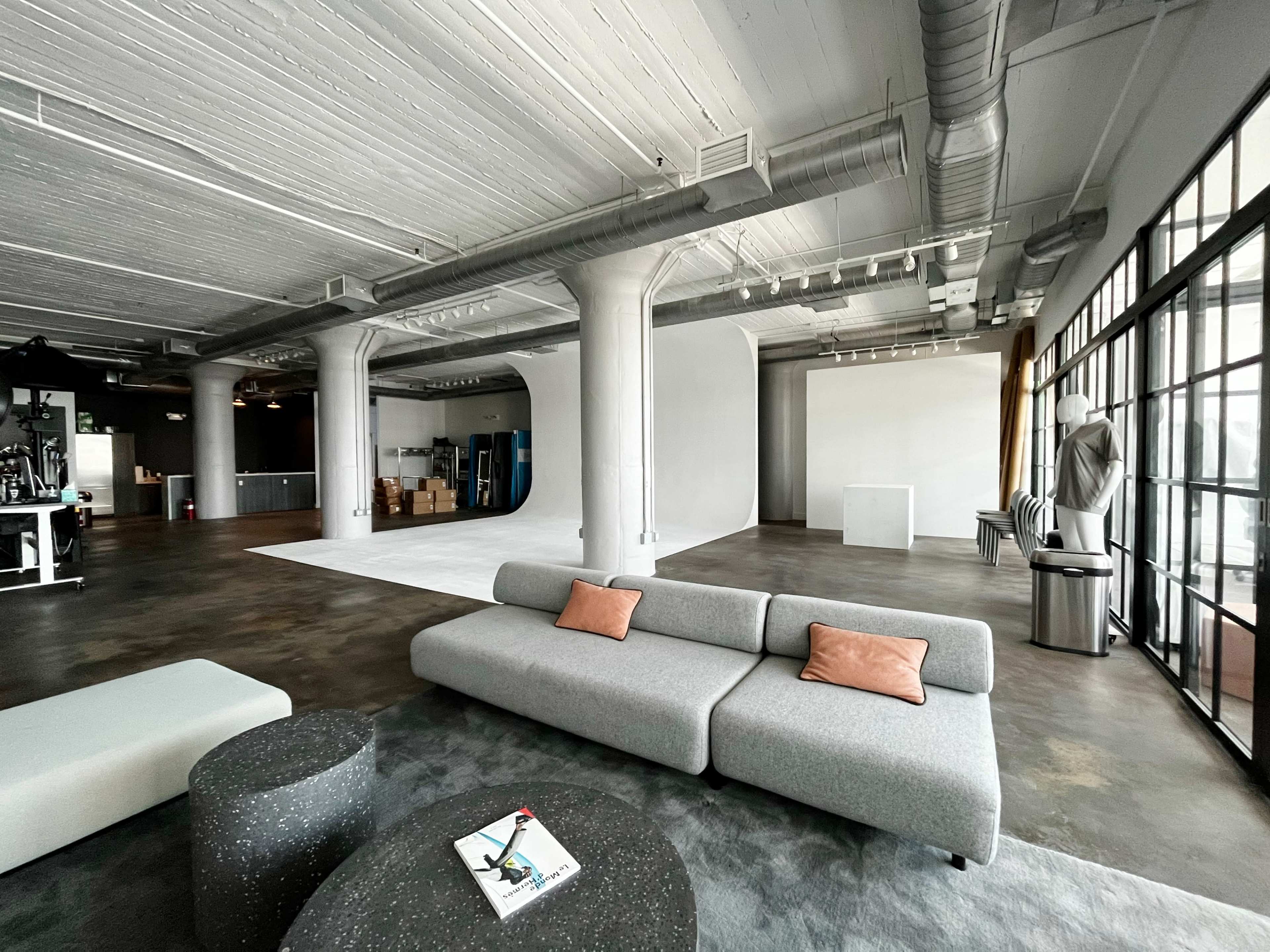 A spacious, industrial-style office interior featuring a gray sofa with two cushions, a round black coffee table, and large windows allowing natural light.