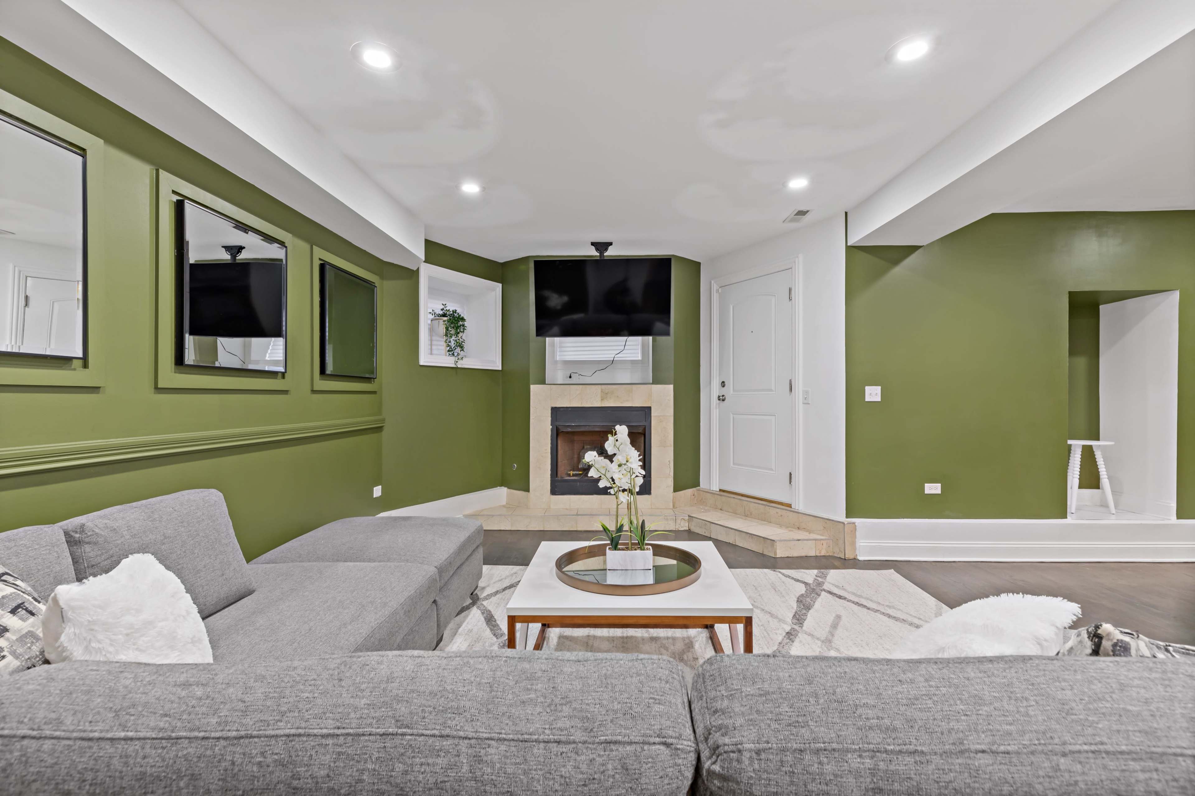 The room features a modern living area with a grey sectional sofa, a fireplace, a mounted television, and green walls adorned with mirrors.