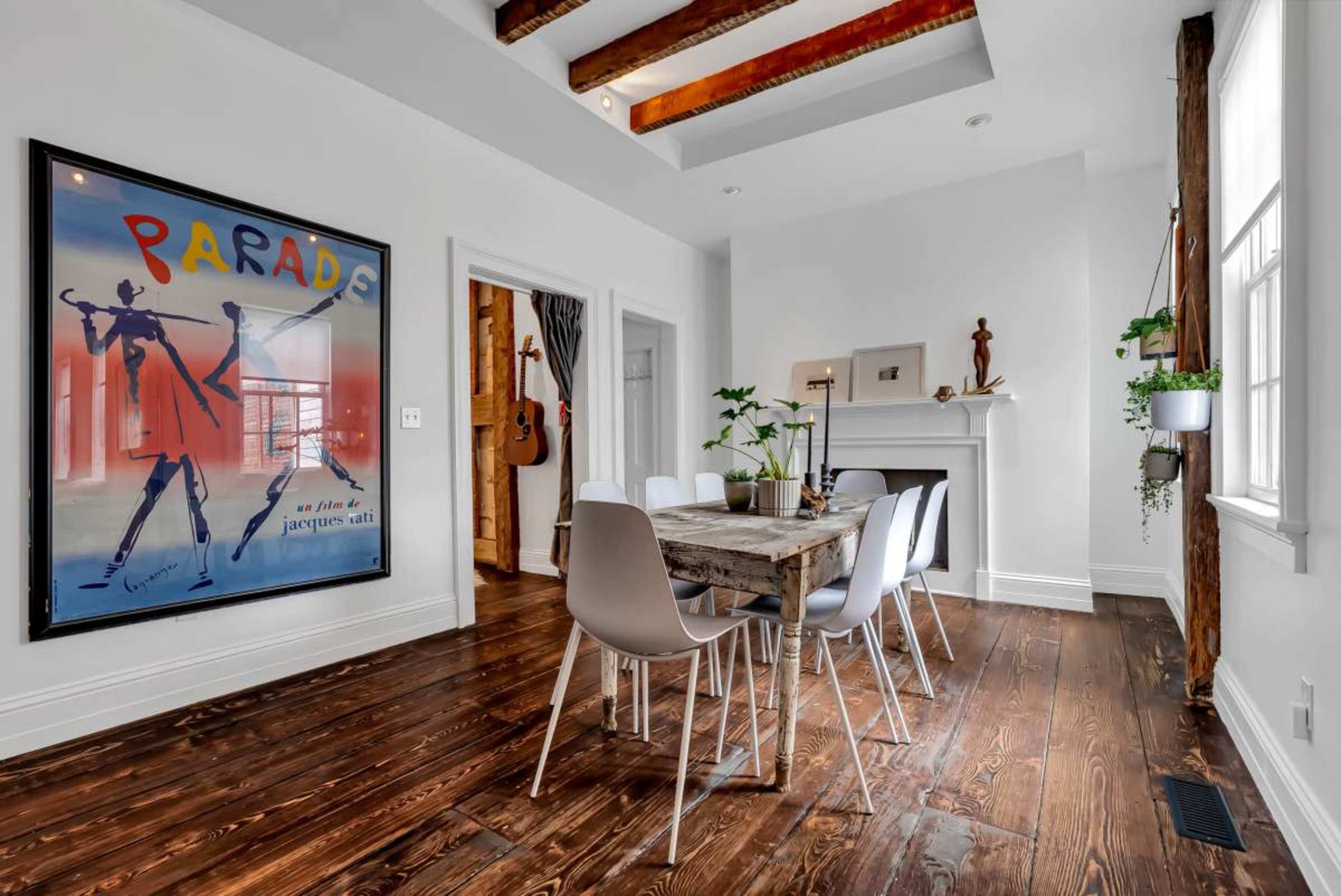 The image displays a modern dining room featuring a wooden table surrounded by white chairs, with a large framed art piece on the wall and a fireplace in the background.