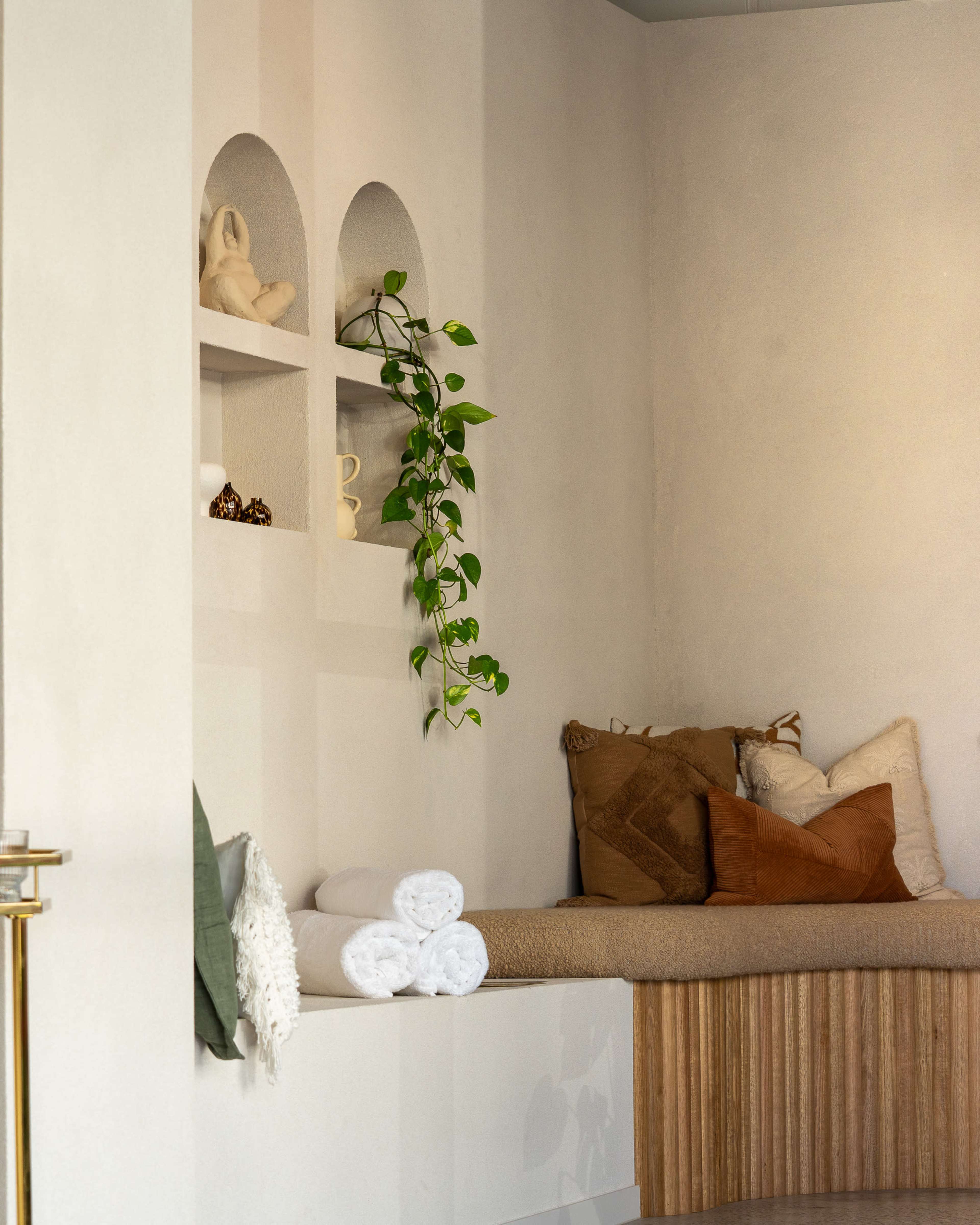 The image features a corner of a room with a bench seat, decorative shelves containing items, and a trailing plant.