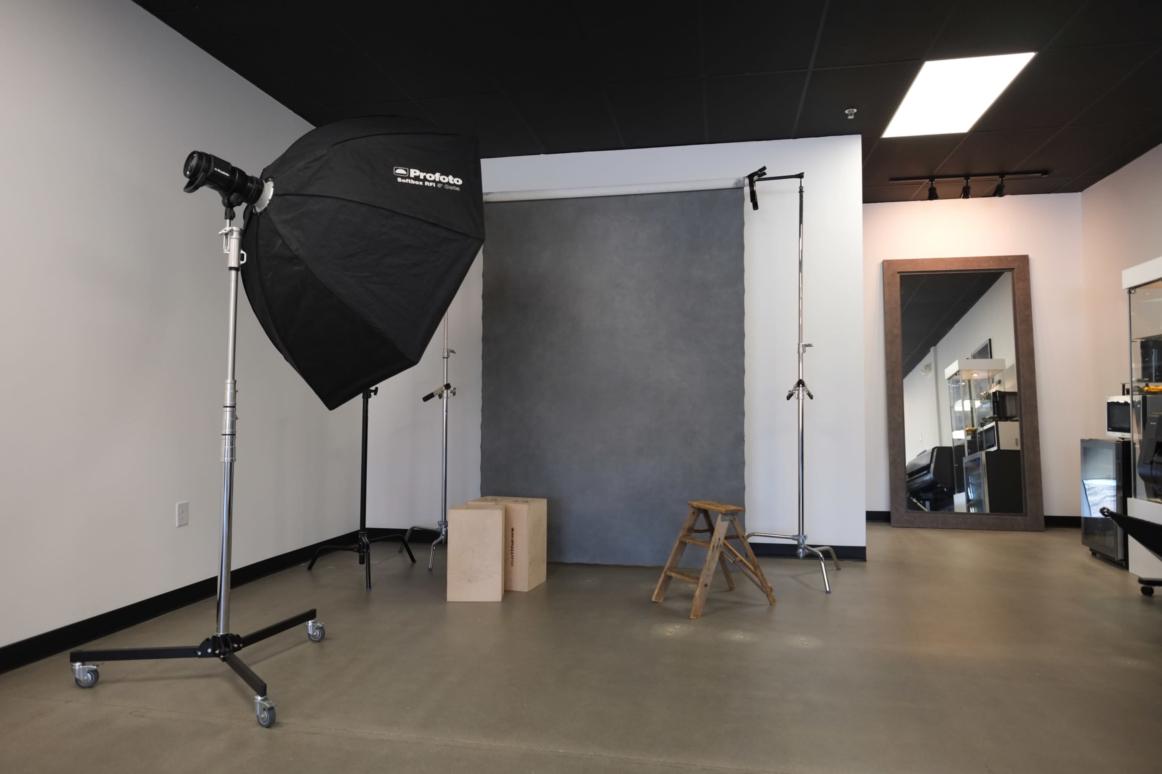 The image shows a photography studio setup with a softbox light, a grey backdrop, and a wooden stool placed in front of it.