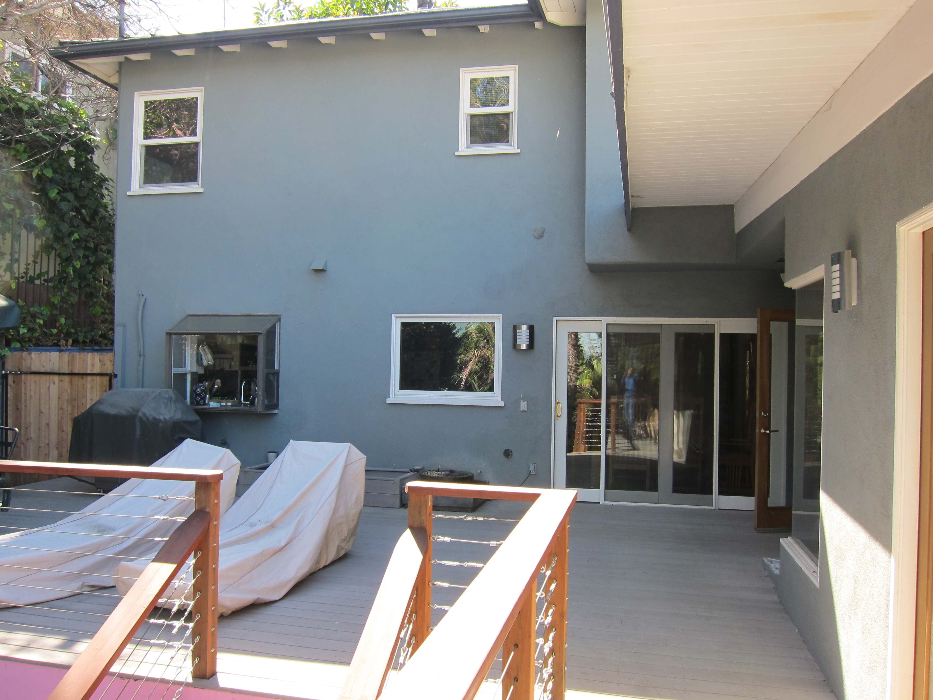 A modern house exterior featuring a wooden deck, covered furniture, and large glass doors leading into the interior.