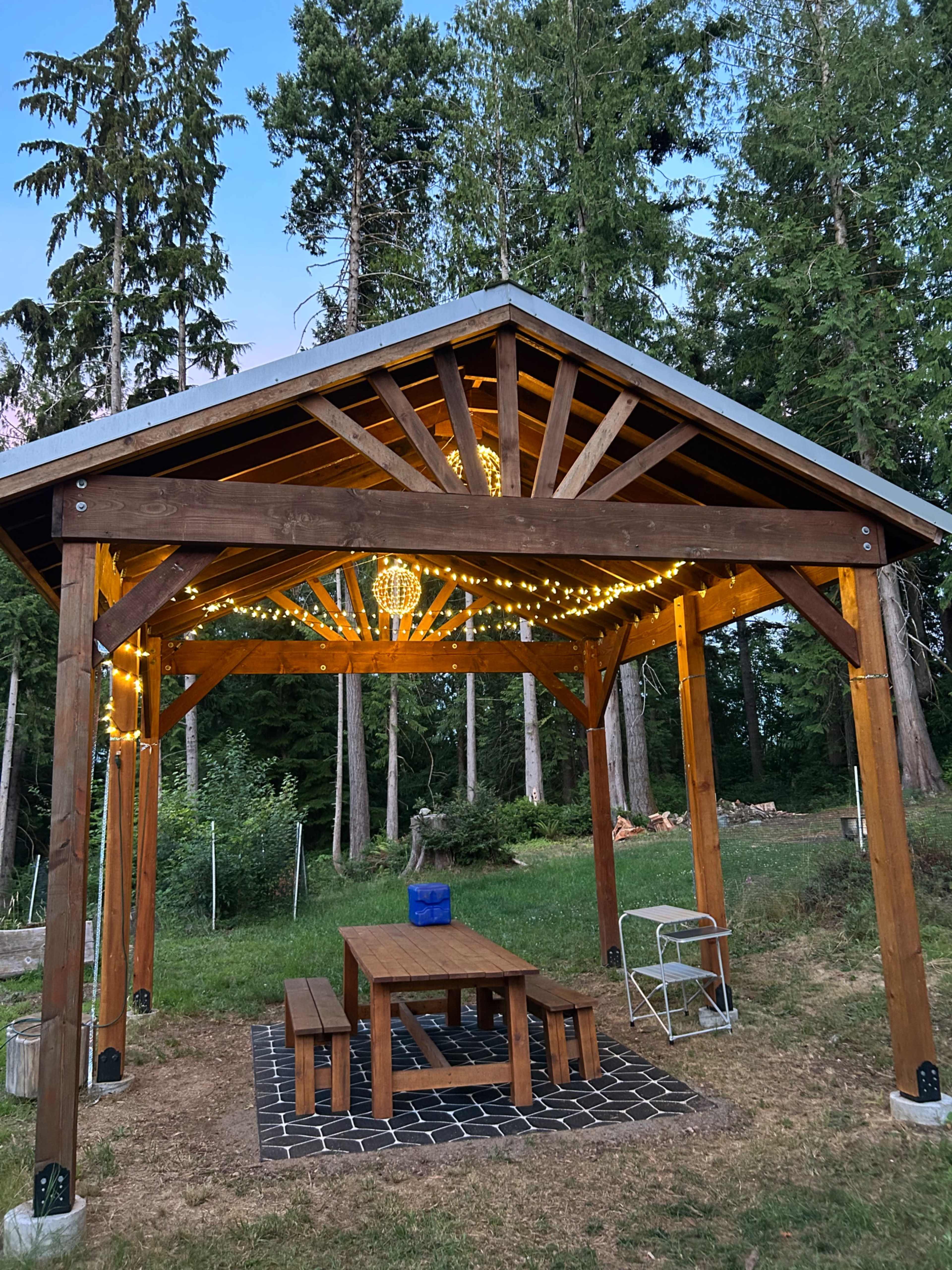 The image shows a wooden pavilion with string lights, featuring a table and benches positioned on a stone patio, surrounded by tall trees.