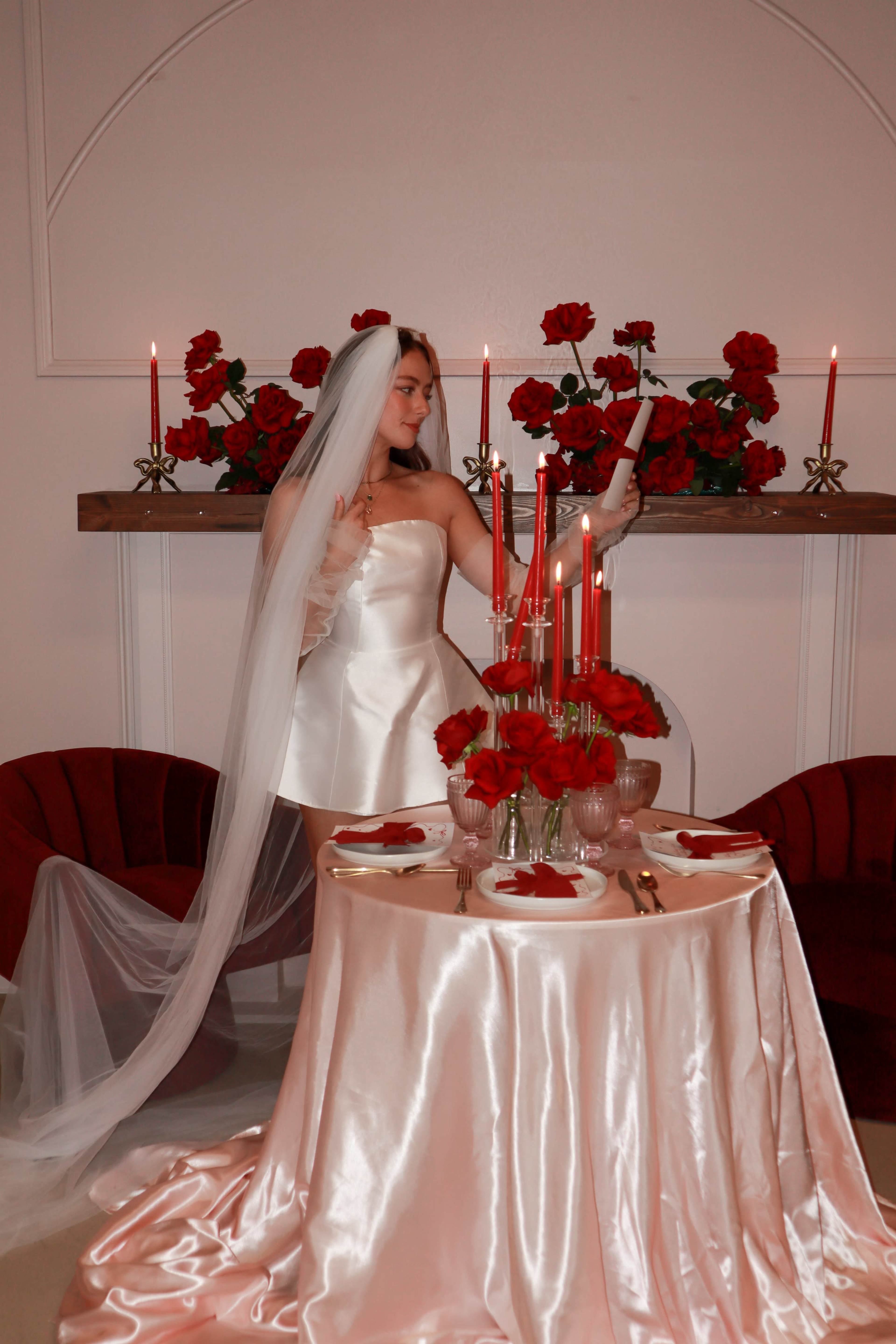 A woman in a satin dress and veil stands by a elegantly set table adorned with red roses and lit candles.