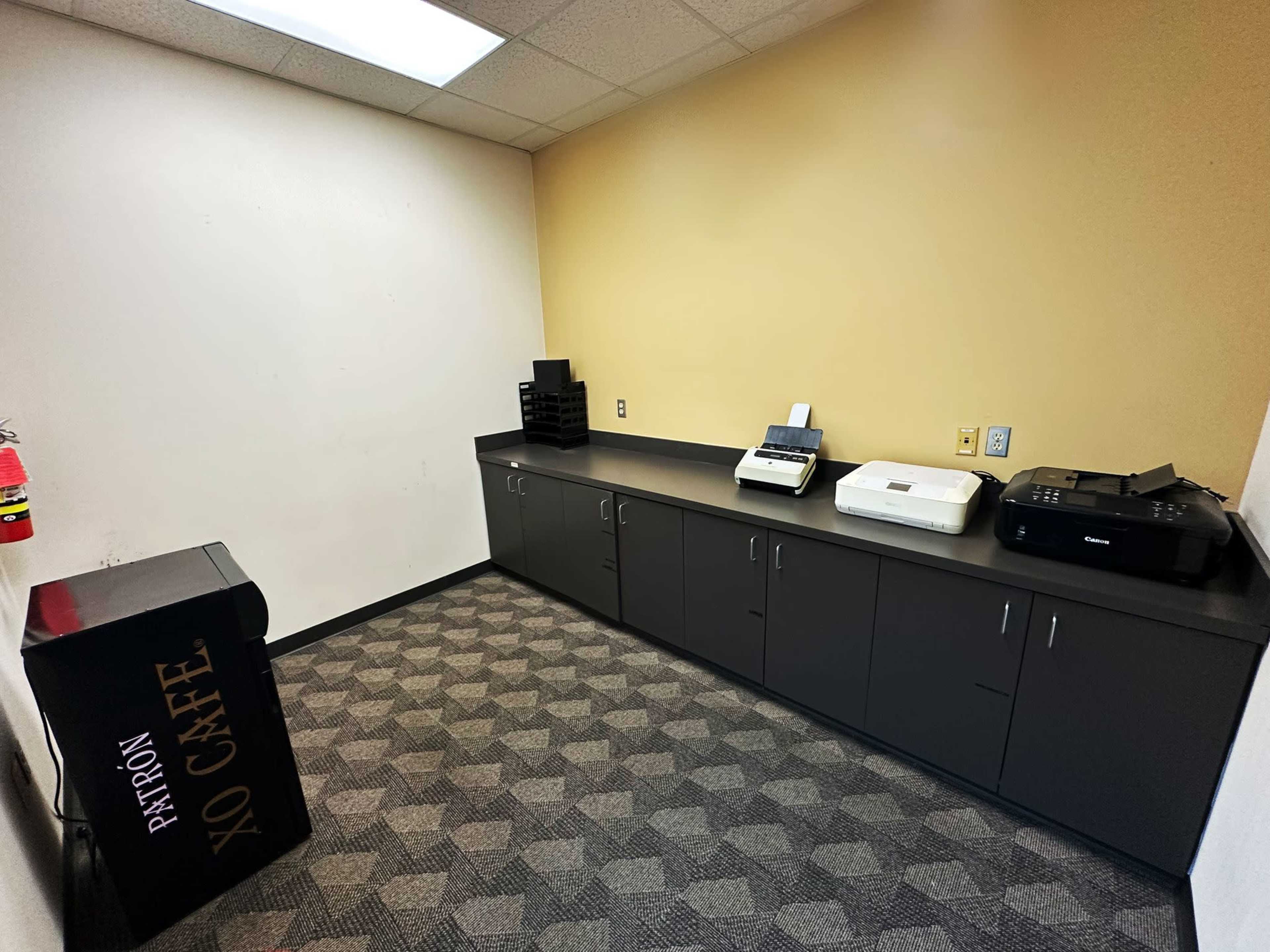 The image shows a small, empty office space with a countertop, two printers, and a black filing cabinet.