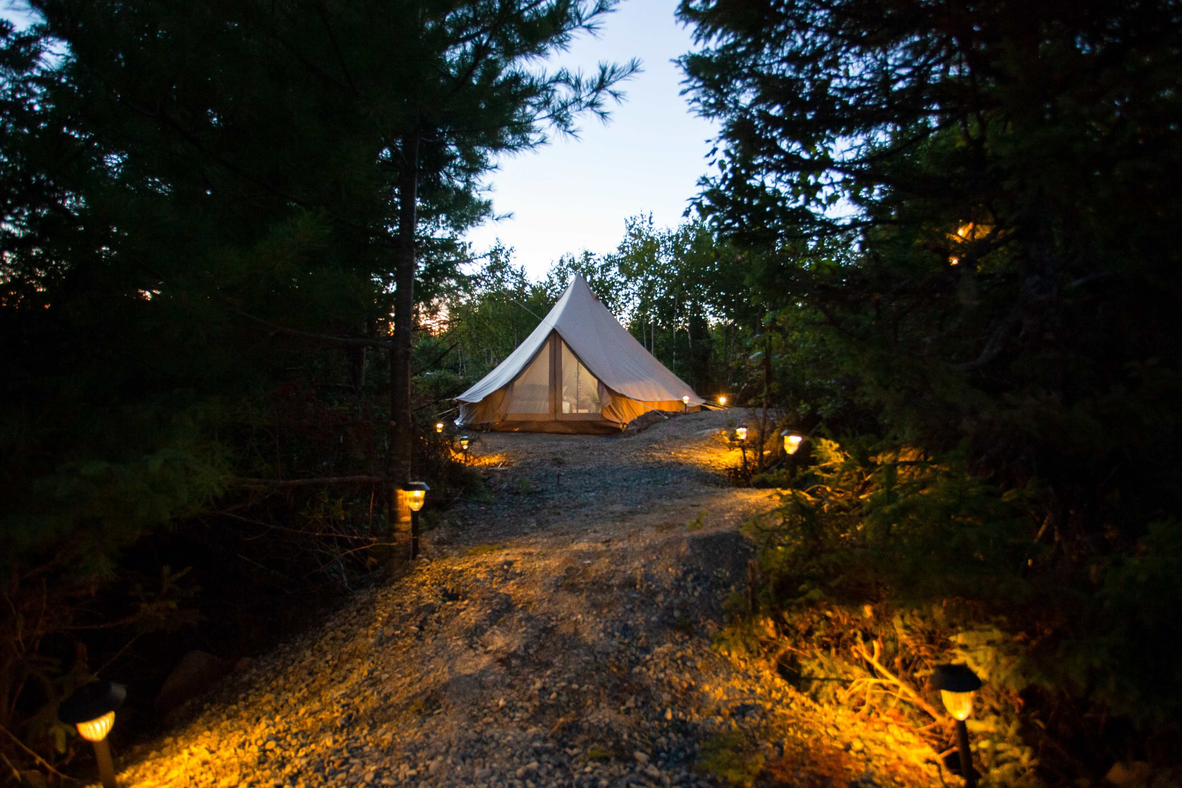 A large canvas tent is positioned at the end of a gravel path, illuminated by ground lights and surrounded by trees.