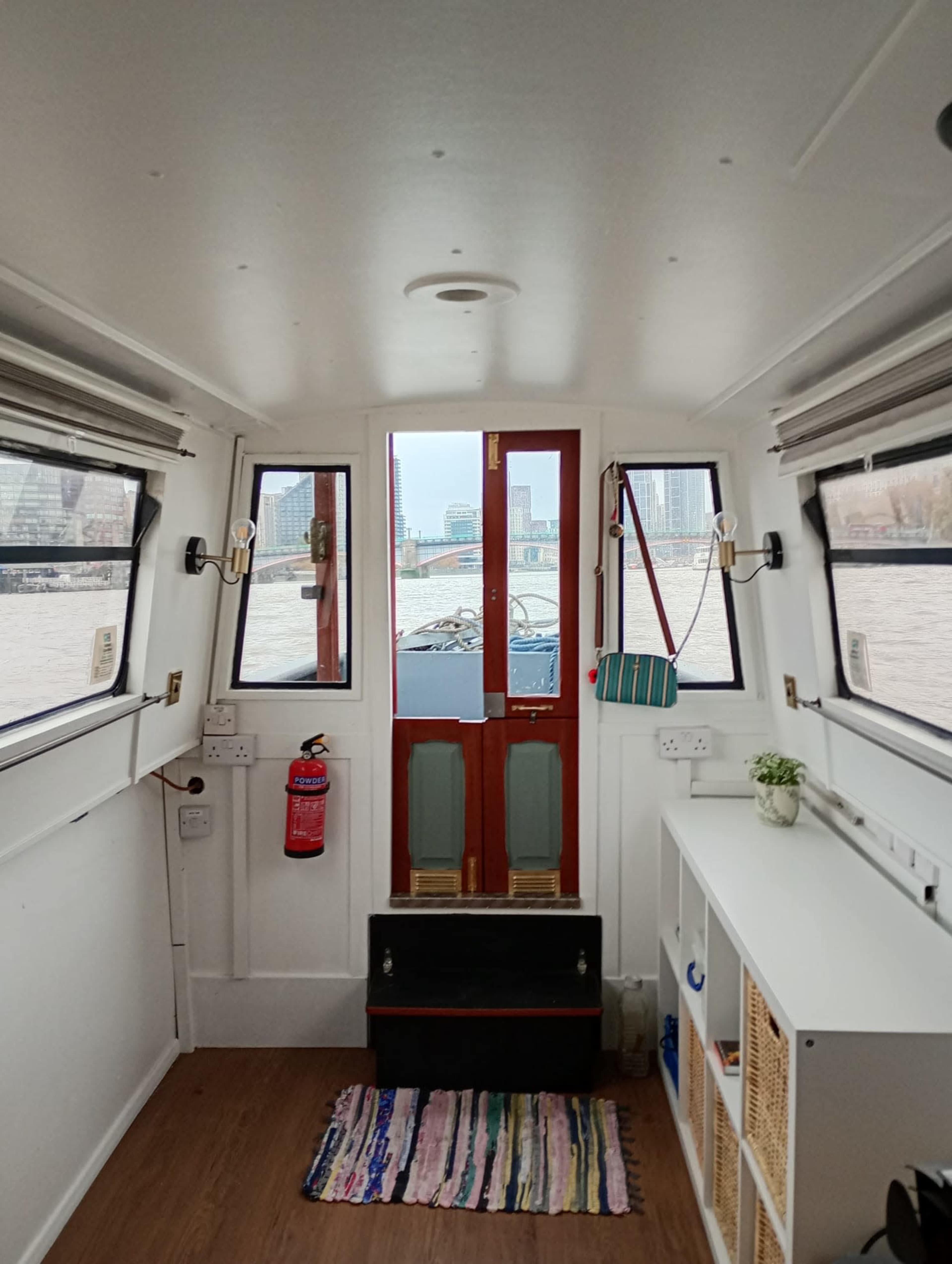 The interior of a narrowboat features a view through the front doors, showcasing the river and urban landscape outside, with a cozy and neatly arranged living space.
