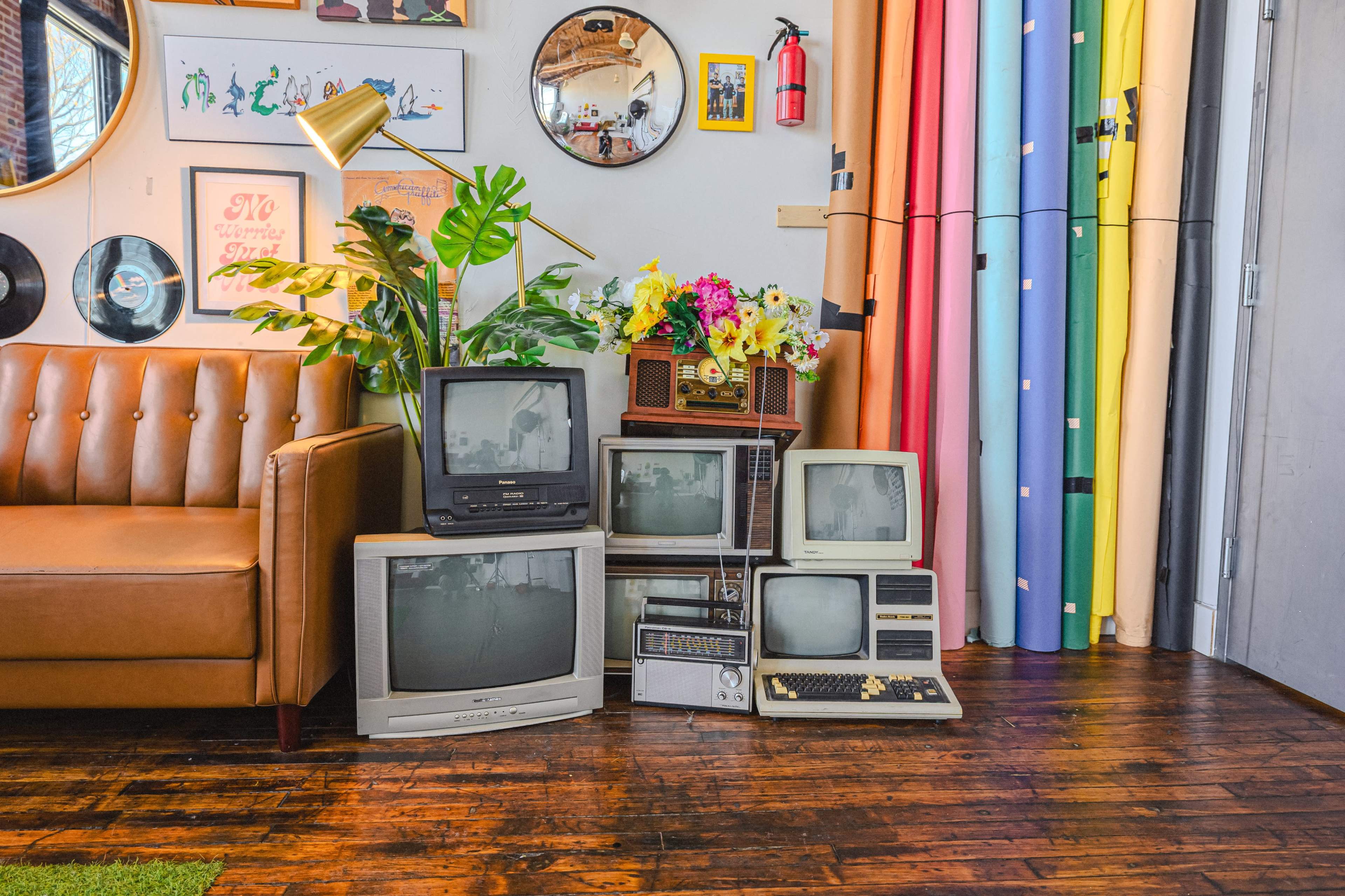 A collection of vintage televisions and a computer are arranged beside a leather couch and a backdrop of colorful rolls of paper in a well-decorated room.