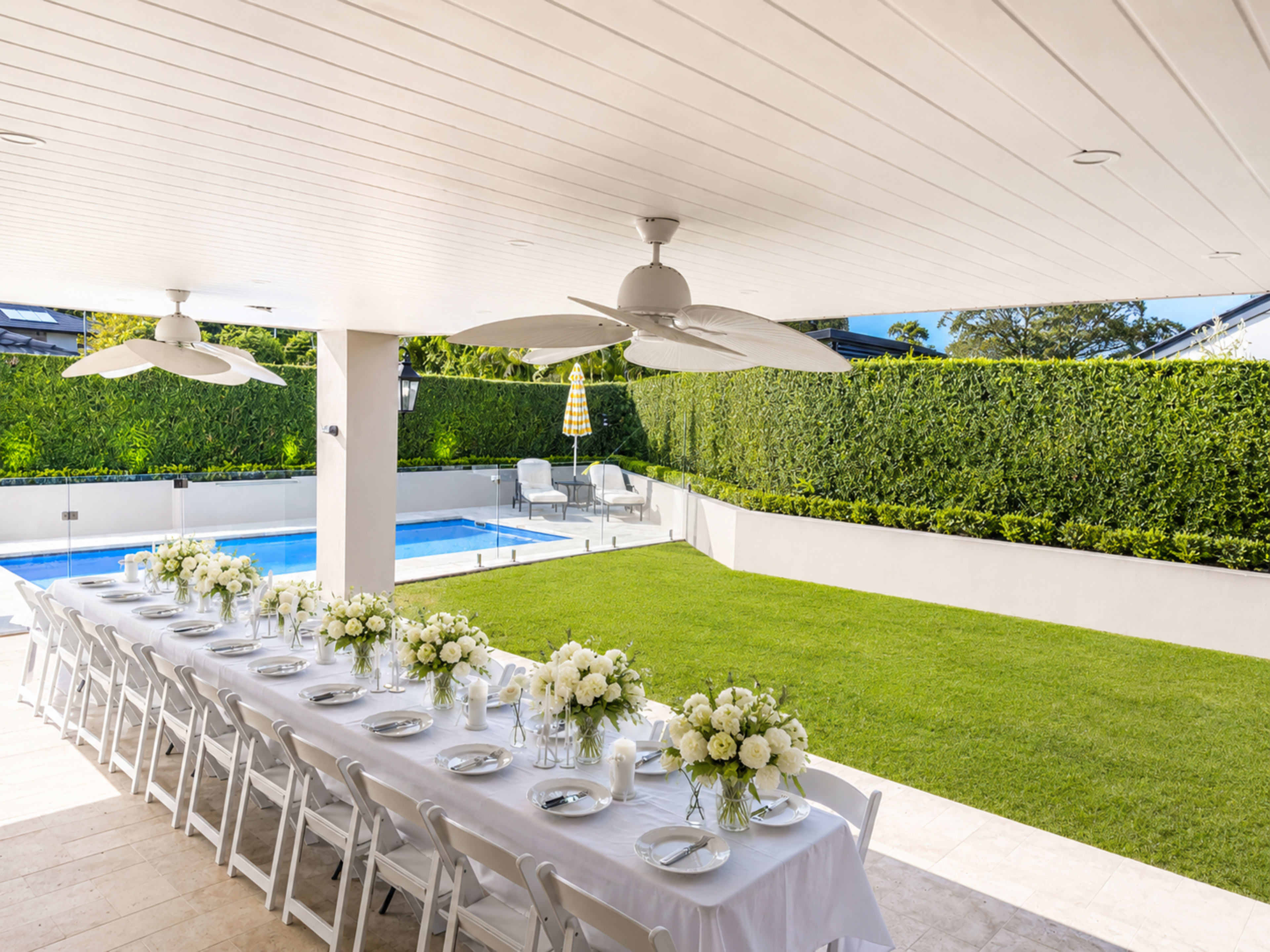 A long dining table adorned with white flowers is set up on a patio beside a swimming pool and a well-maintained lawn.
