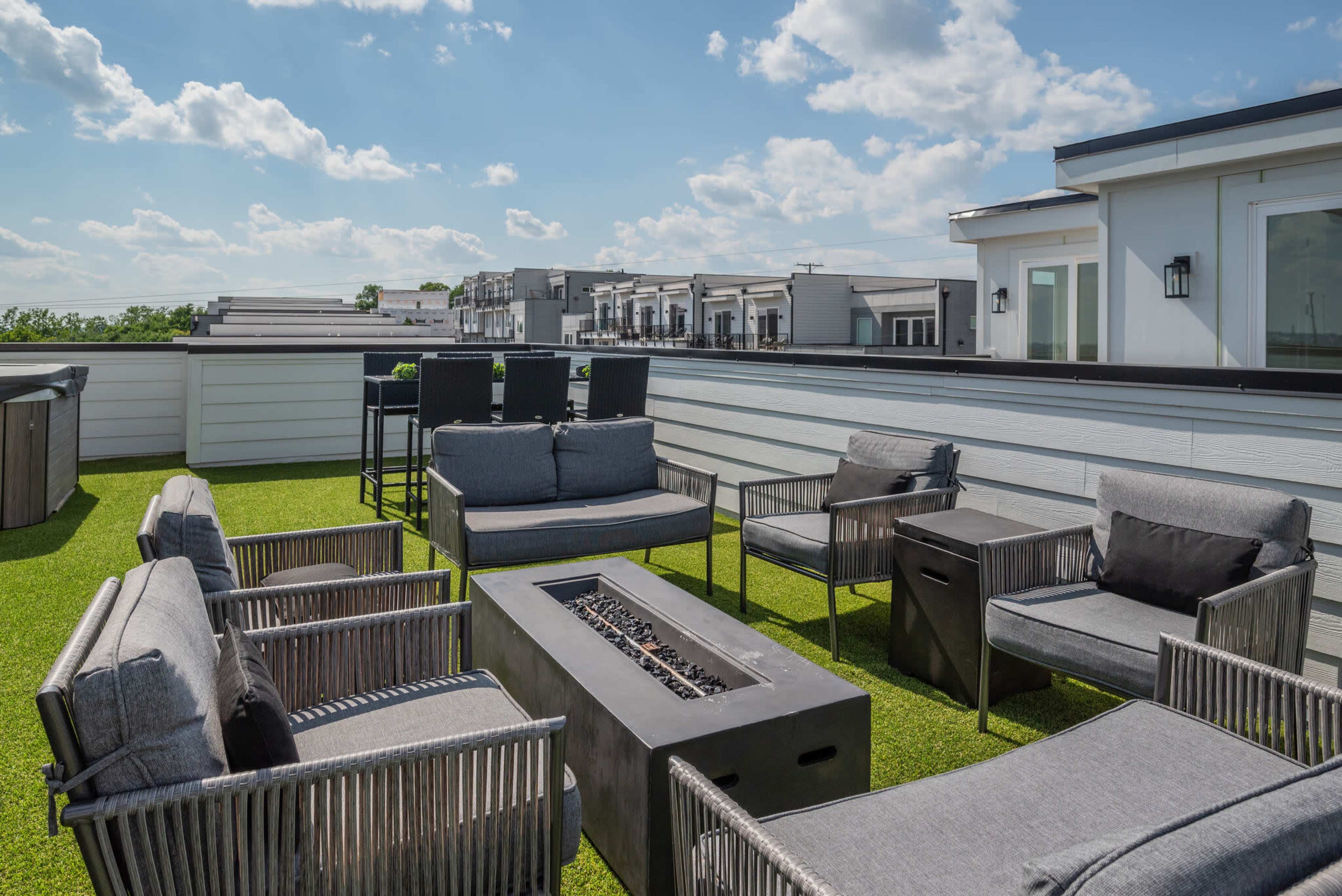 A rooftop terrace features gray furniture arranged around a fire pit, with green synthetic grass and a backdrop of buildings under a partly cloudy sky.
