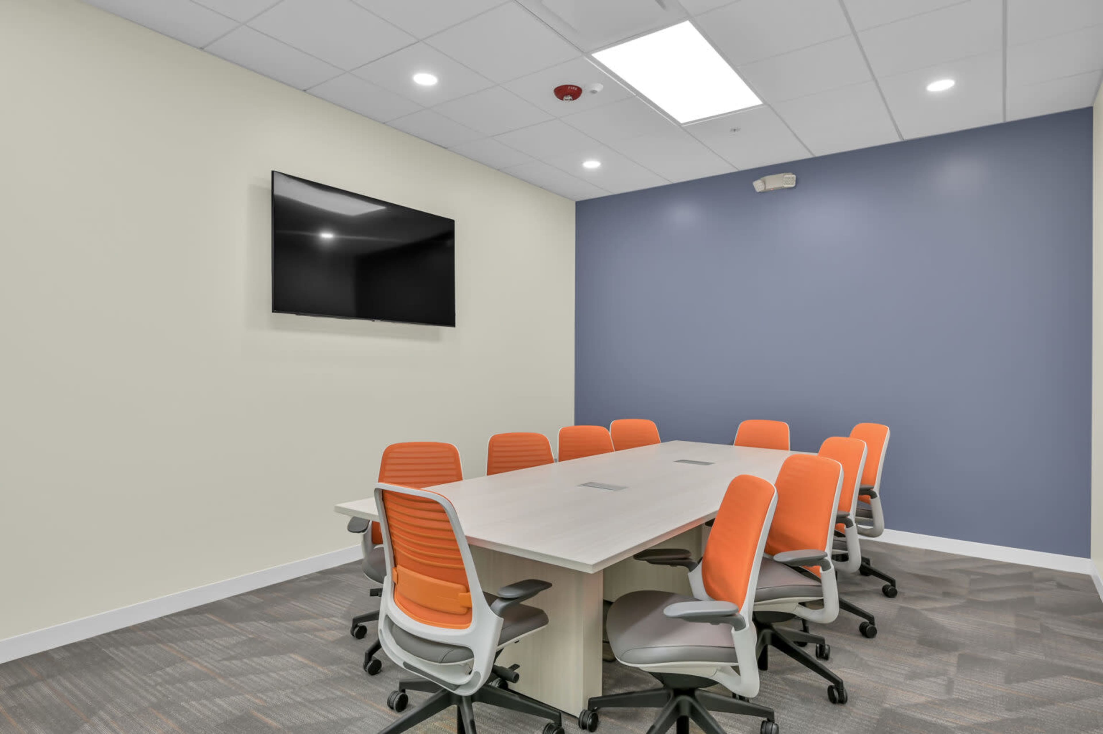 A modern conference room features a large table surrounded by eight orange and gray chairs, with a wall-mounted television and light-colored walls.