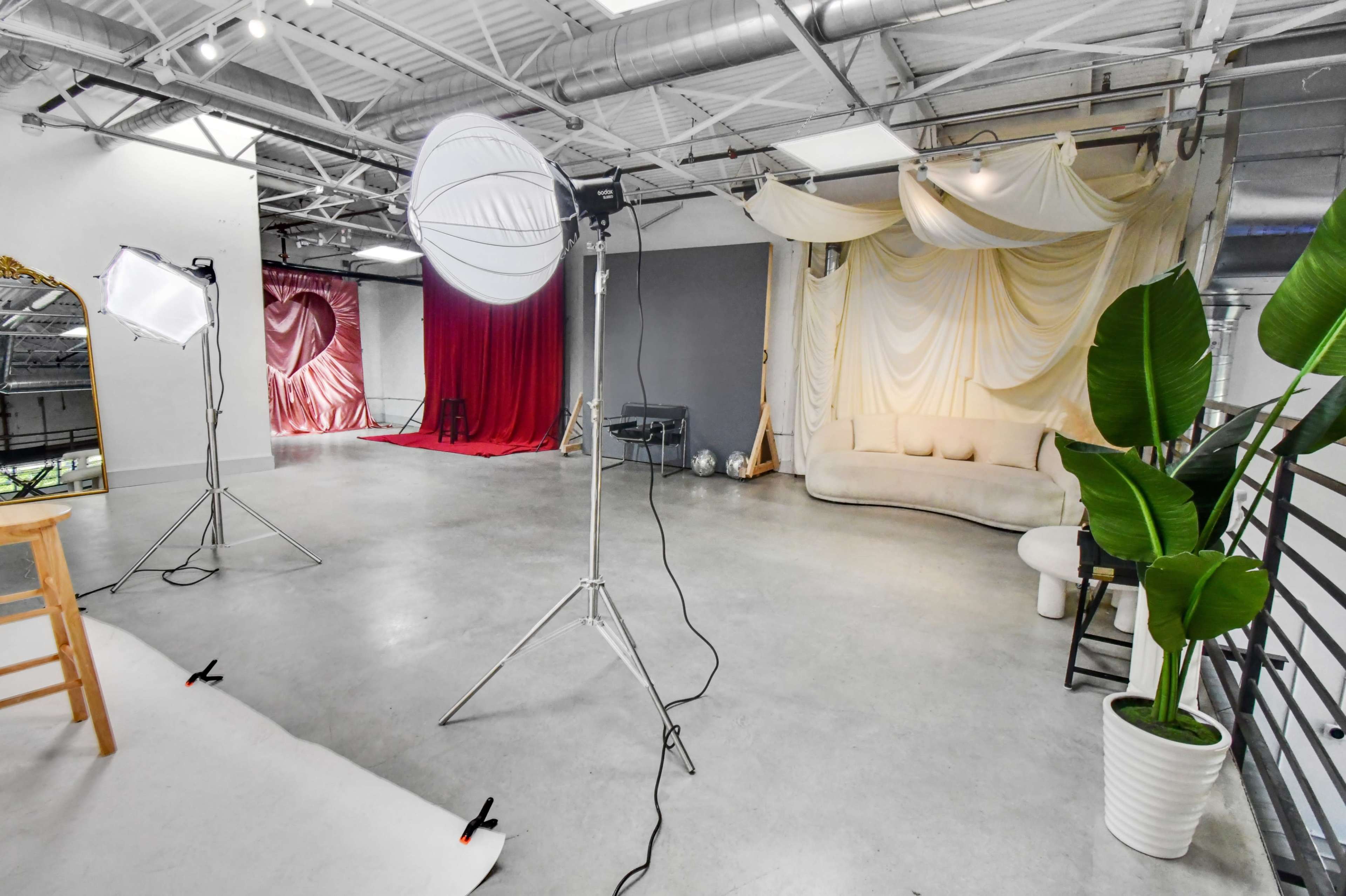 The image shows a spacious photography studio featuring soft lighting equipment, a white backdrop, red and beige fabric drapes, and various seating arrangements.