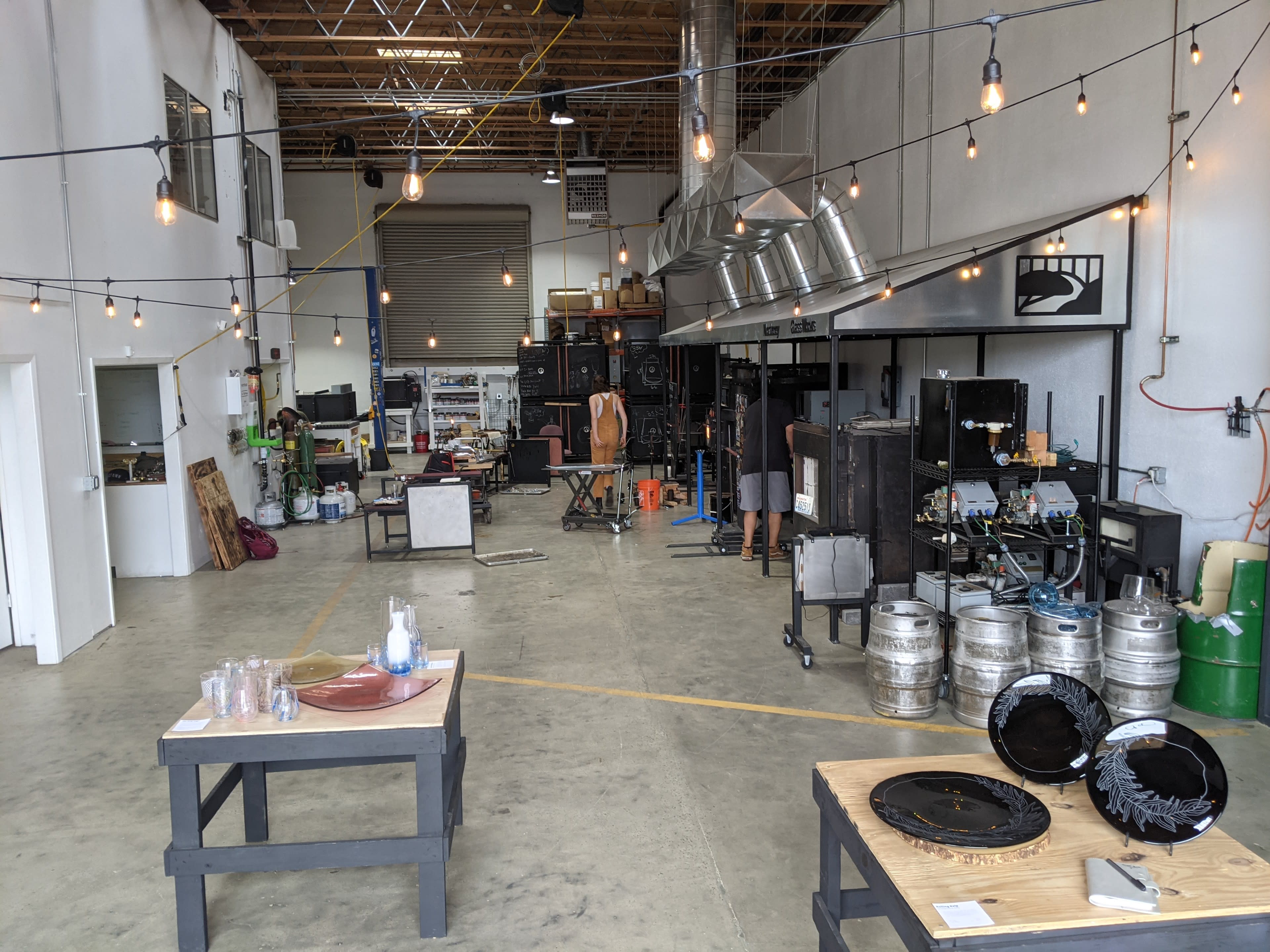 A spacious workshop with various equipment and tools, illuminated by hanging light bulbs, featuring glassware and metal trays on wooden tables.