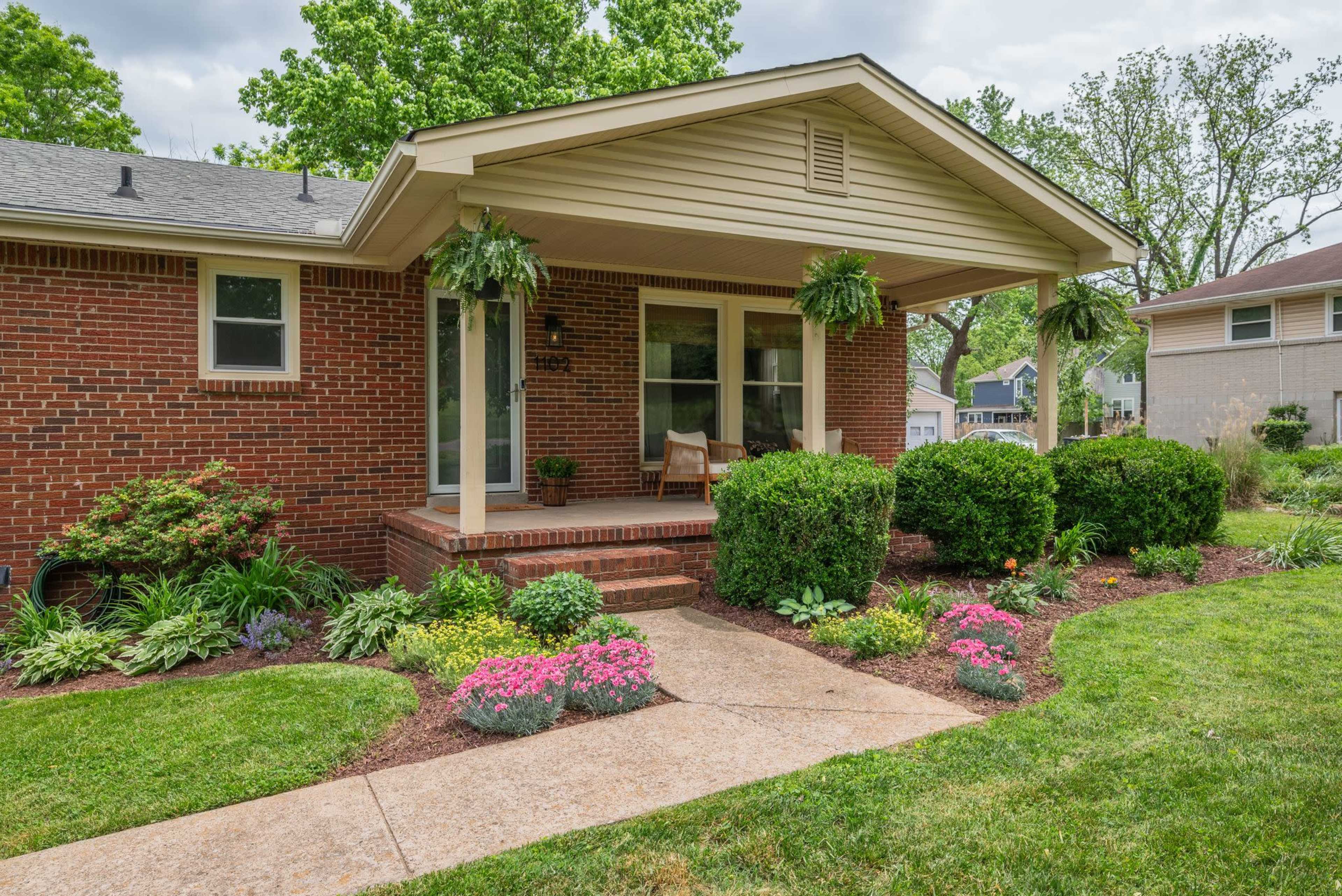 A brick house with a front porch, flanked by decorative plants and flower beds along a walkway.