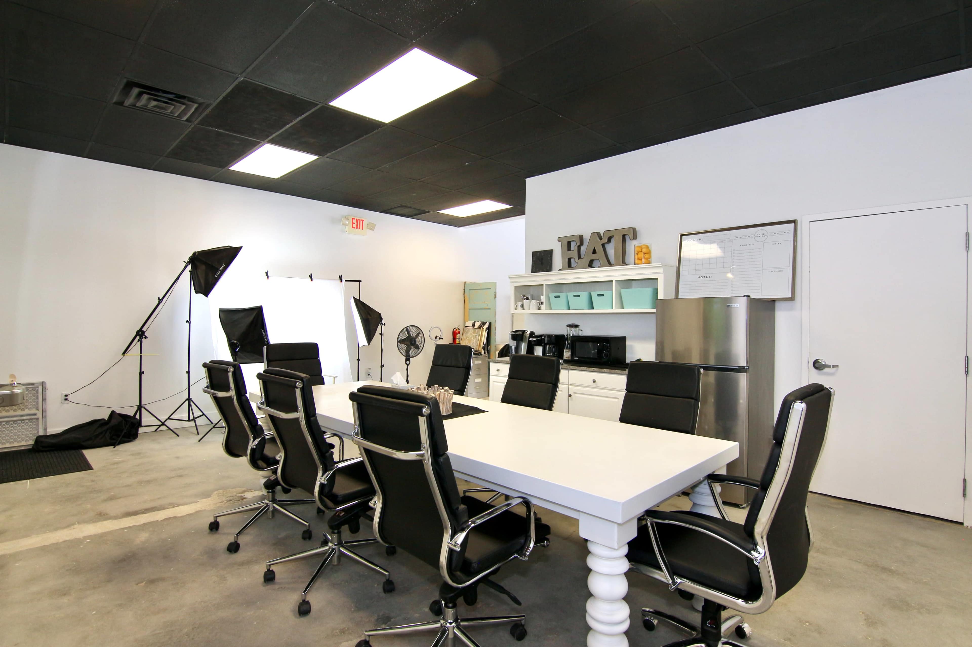 A modern conference room features a long white table surrounded by black chairs, with photography equipment and a kitchenette visible in the background.