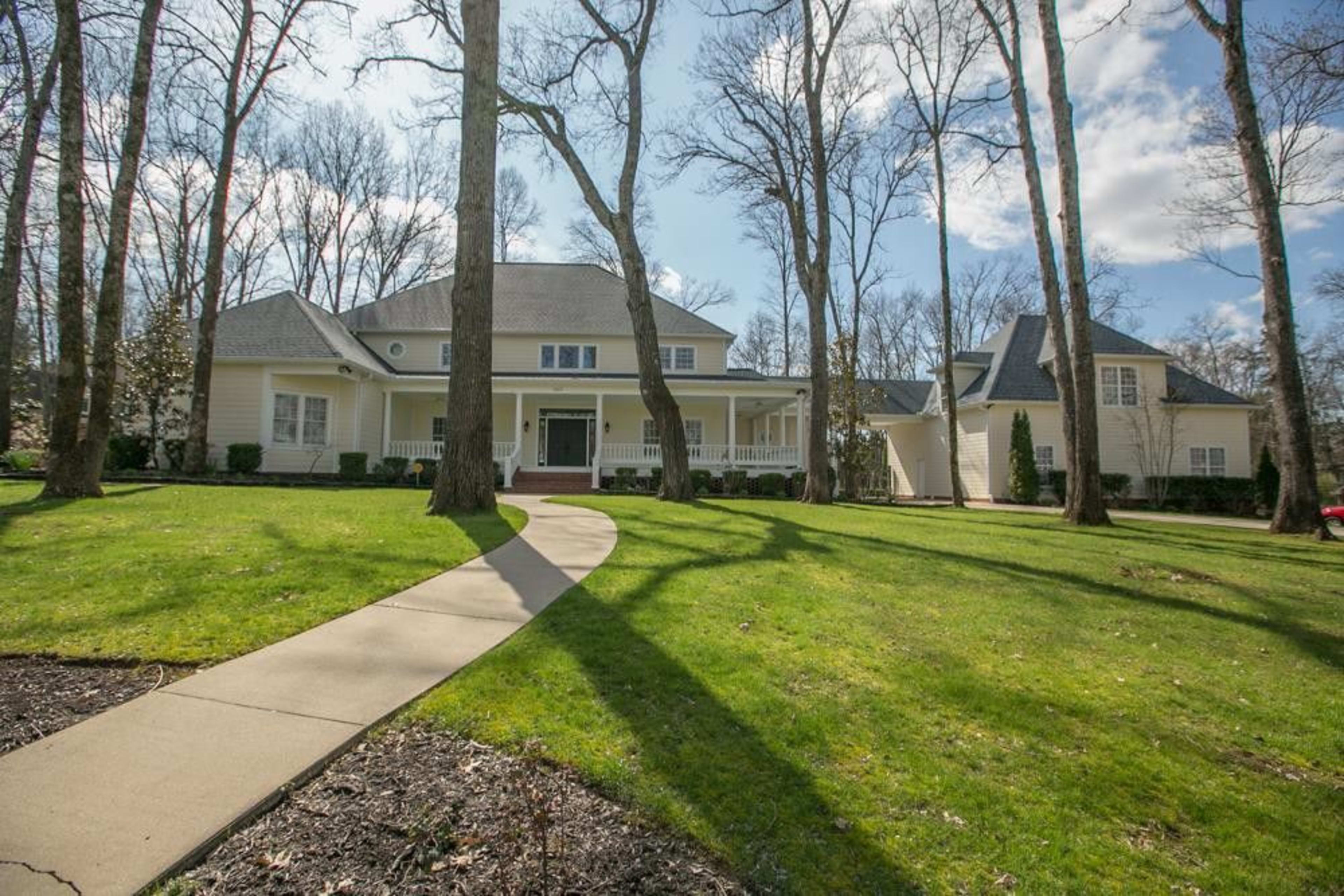 A spacious two-story house is surrounded by tall trees and a manicured lawn, with a winding walkway leading to the entrance.