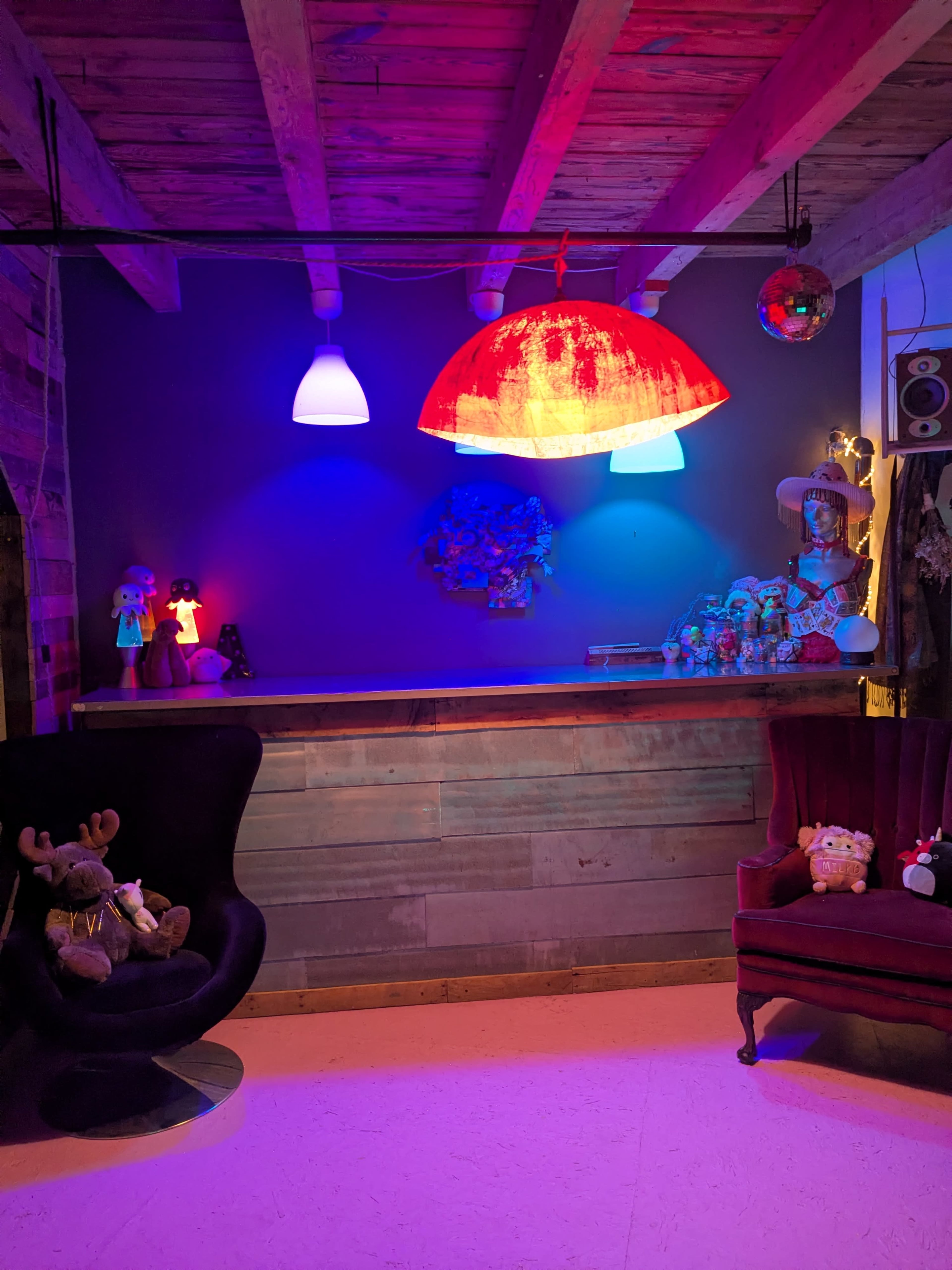The image shows a cozy interior space with a wooden bar, colorful pendant lights, and a mix of plush toys and decorative items on display.