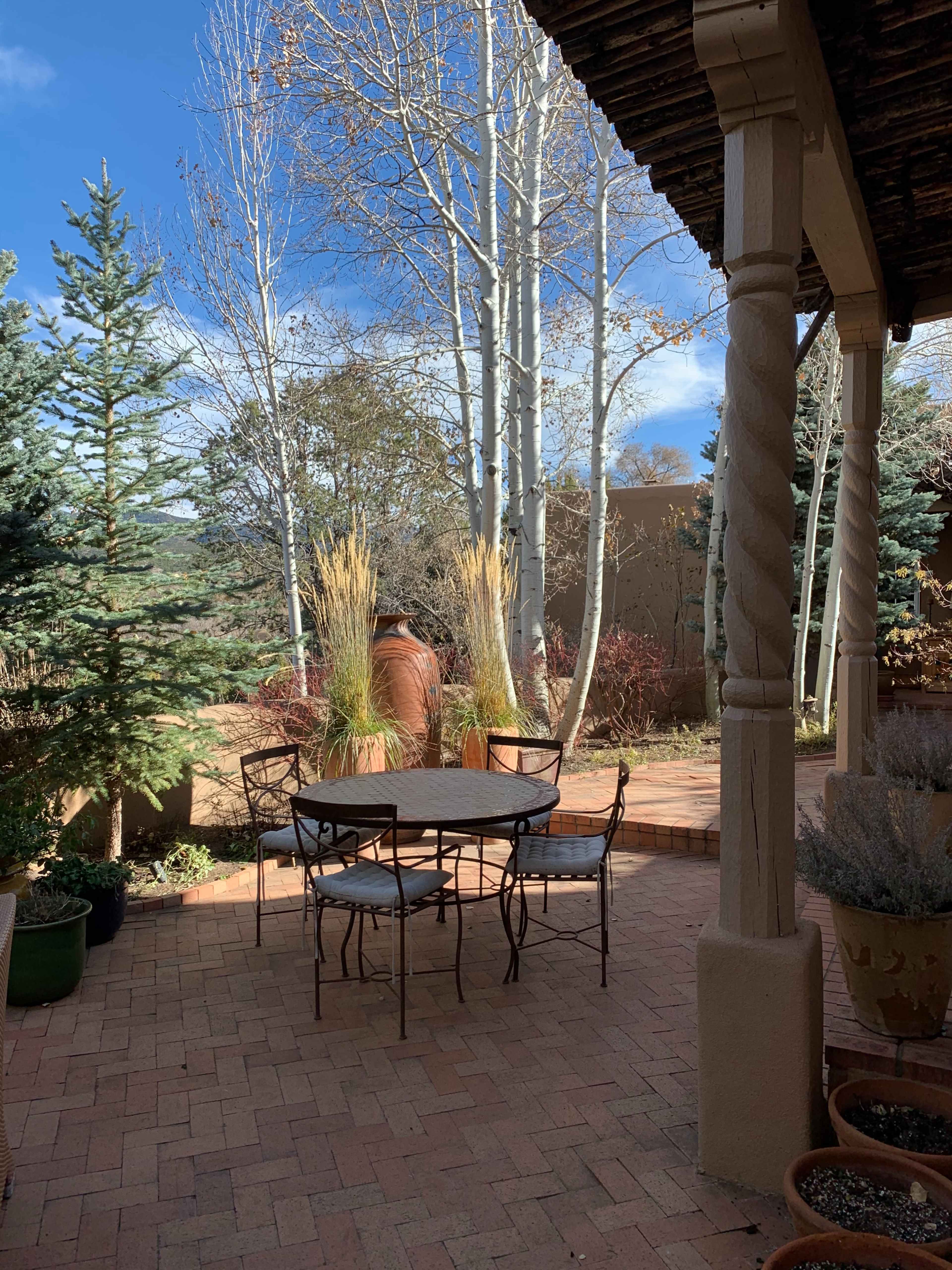 A small round metal table with four chairs is set on a brick patio surrounded by trees and shrubs.