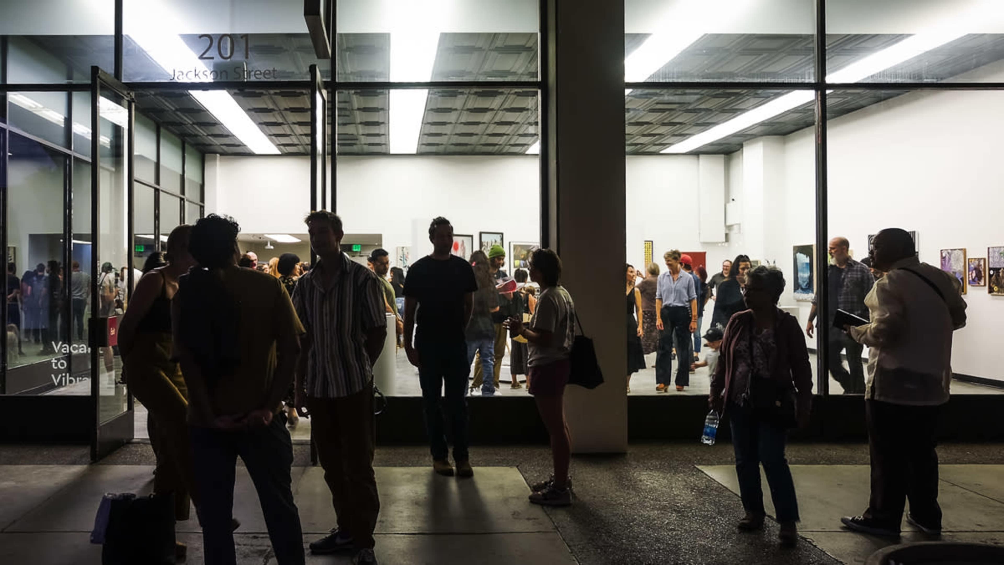 A crowd of people gathers outside an art gallery at night, with a well-lit exhibition visible through large glass windows.