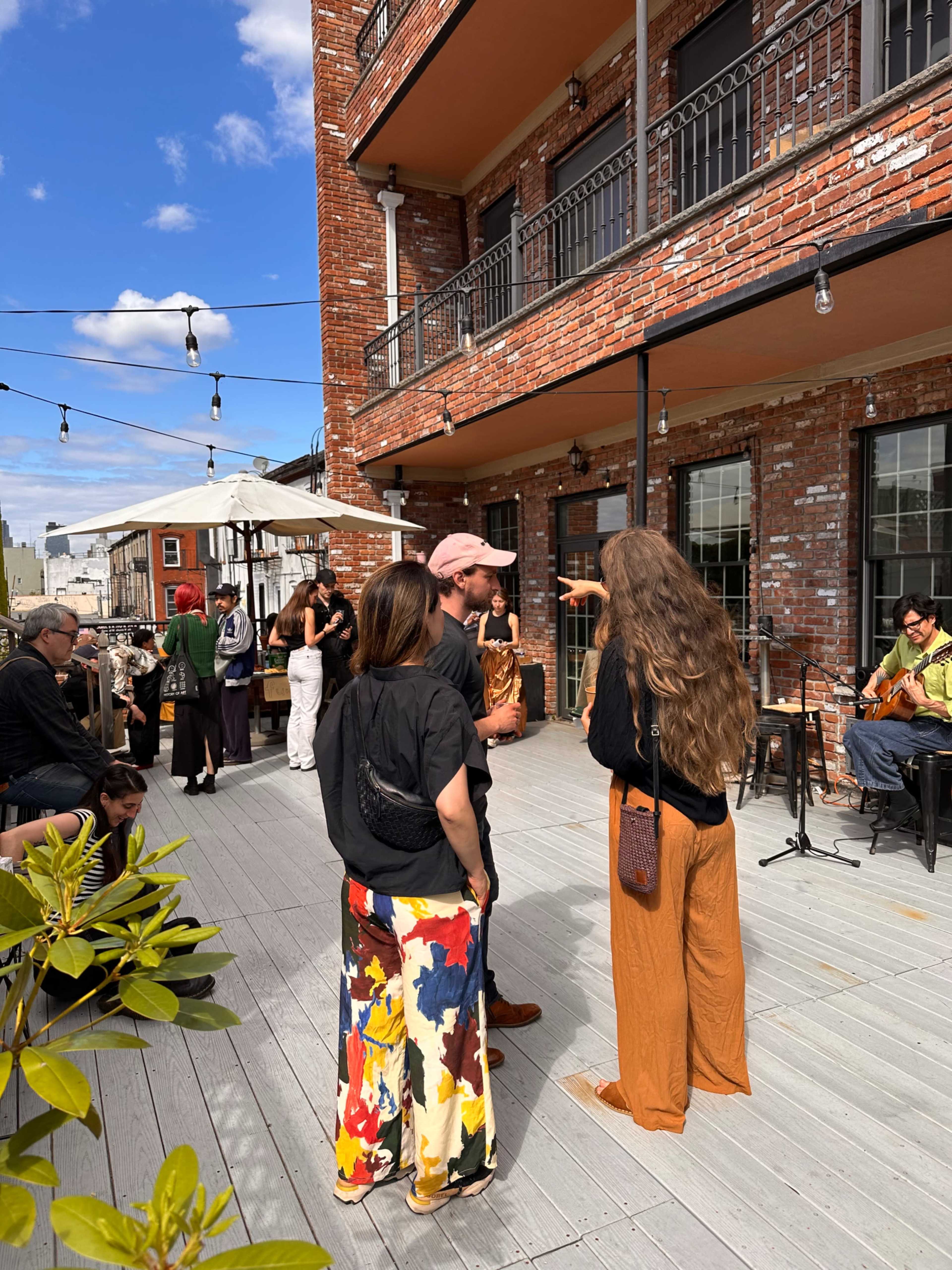 A group of people stands on a deck surrounded by brick buildings while a musician plays in the background.
