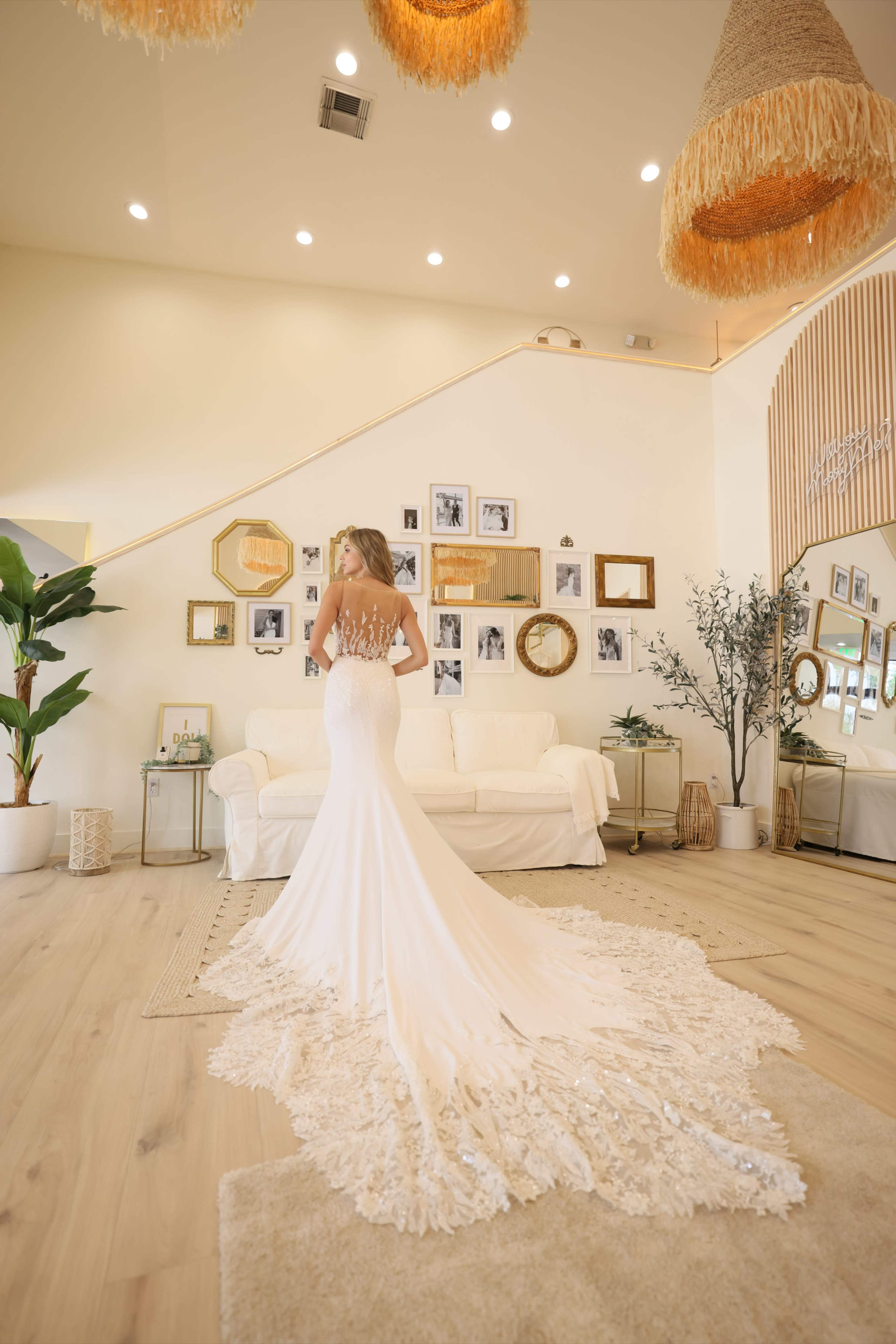 A woman in a wedding dress stands with her back to the camera in a bright, elegantly decorated room featuring various framed photographs on the walls.