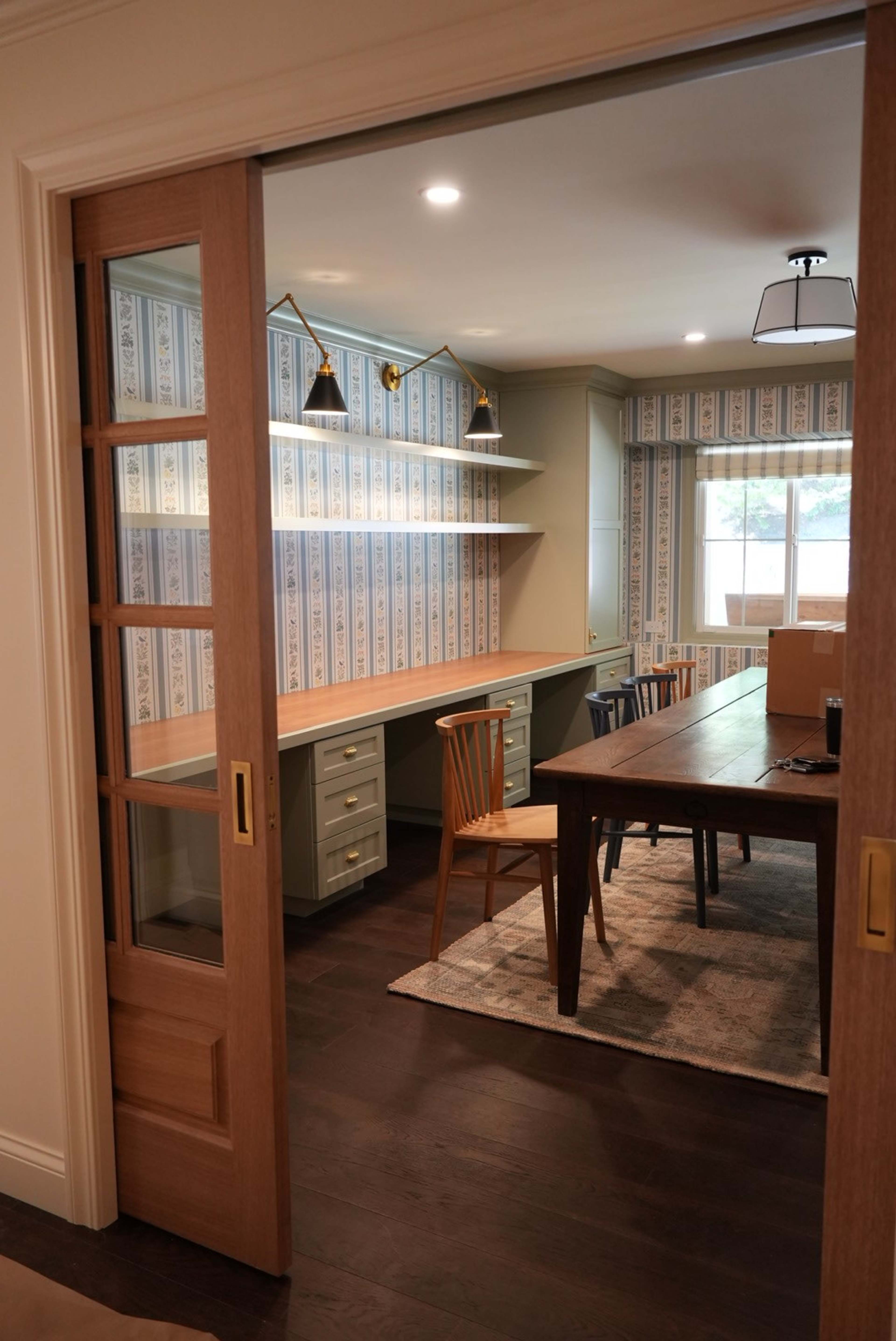 The image shows a room with a wooden dining table and chairs, a built-in desk with shelves, and patterned wallpaper, viewed through an open sliding door.