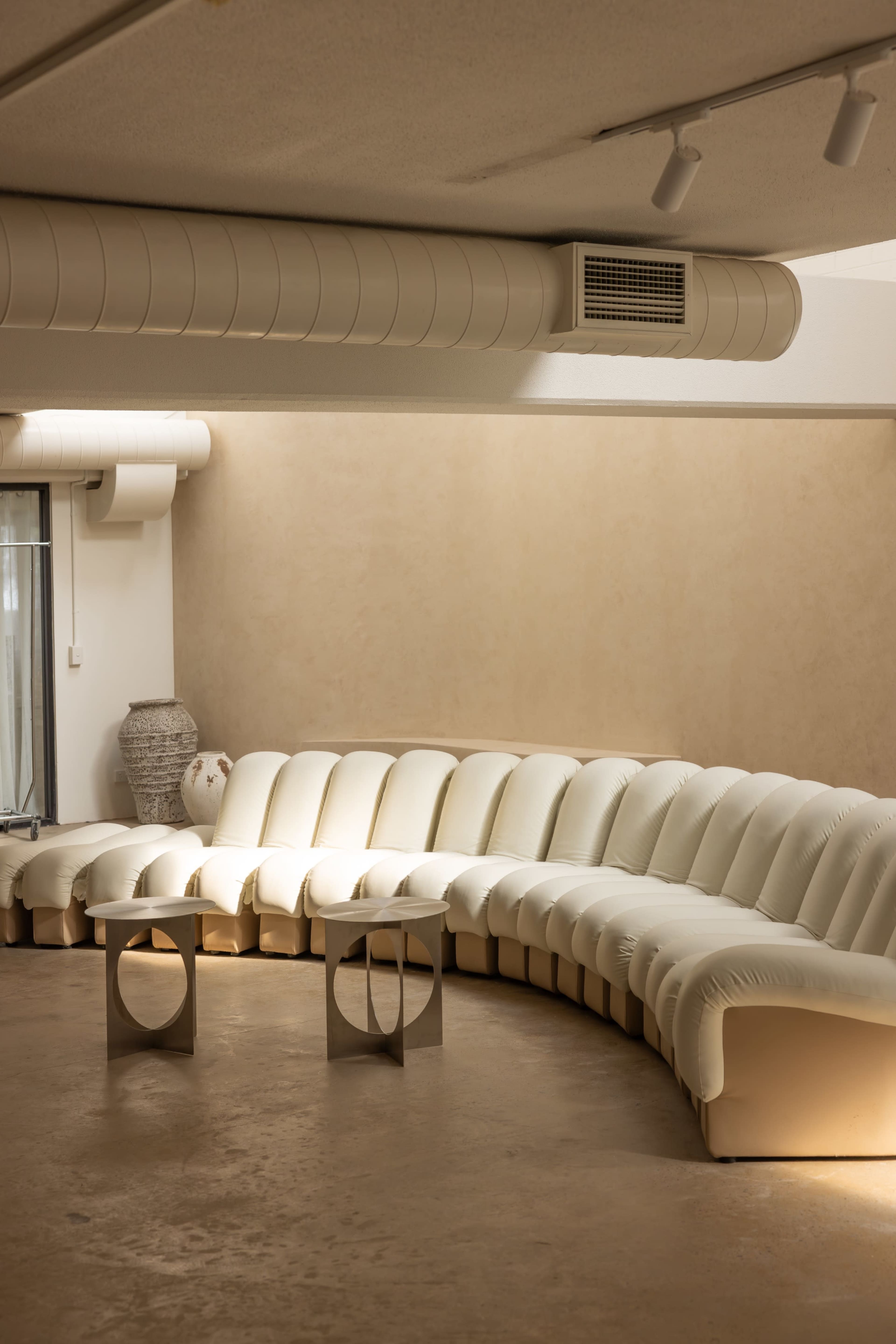 The image shows a curved, white upholstered sofa arranged in a circular formation with two metallic tables in front of it in a minimalist interior space.
