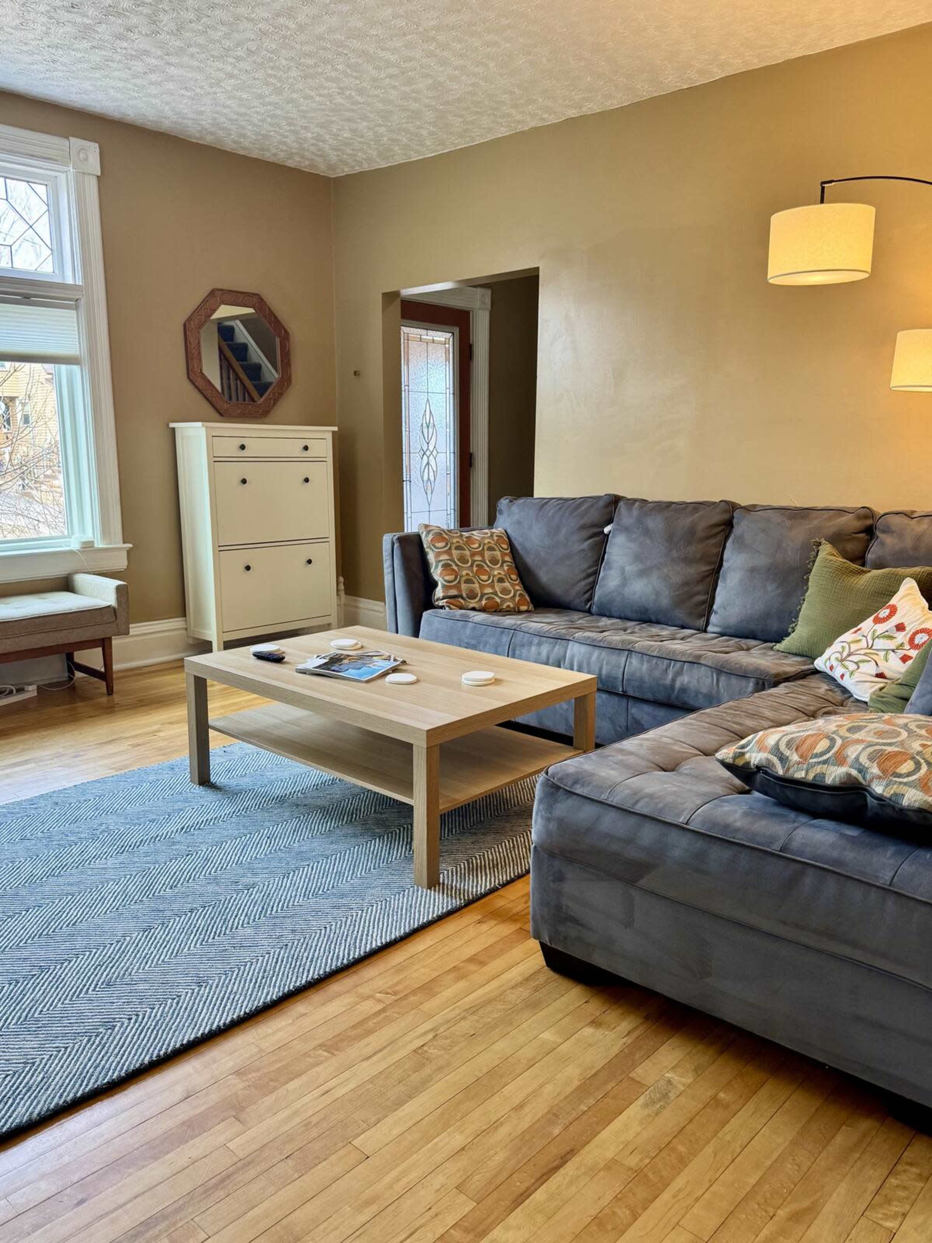 The image shows a living room with a gray sectional couch, a wooden coffee table, and a light-colored area rug on hardwood flooring.