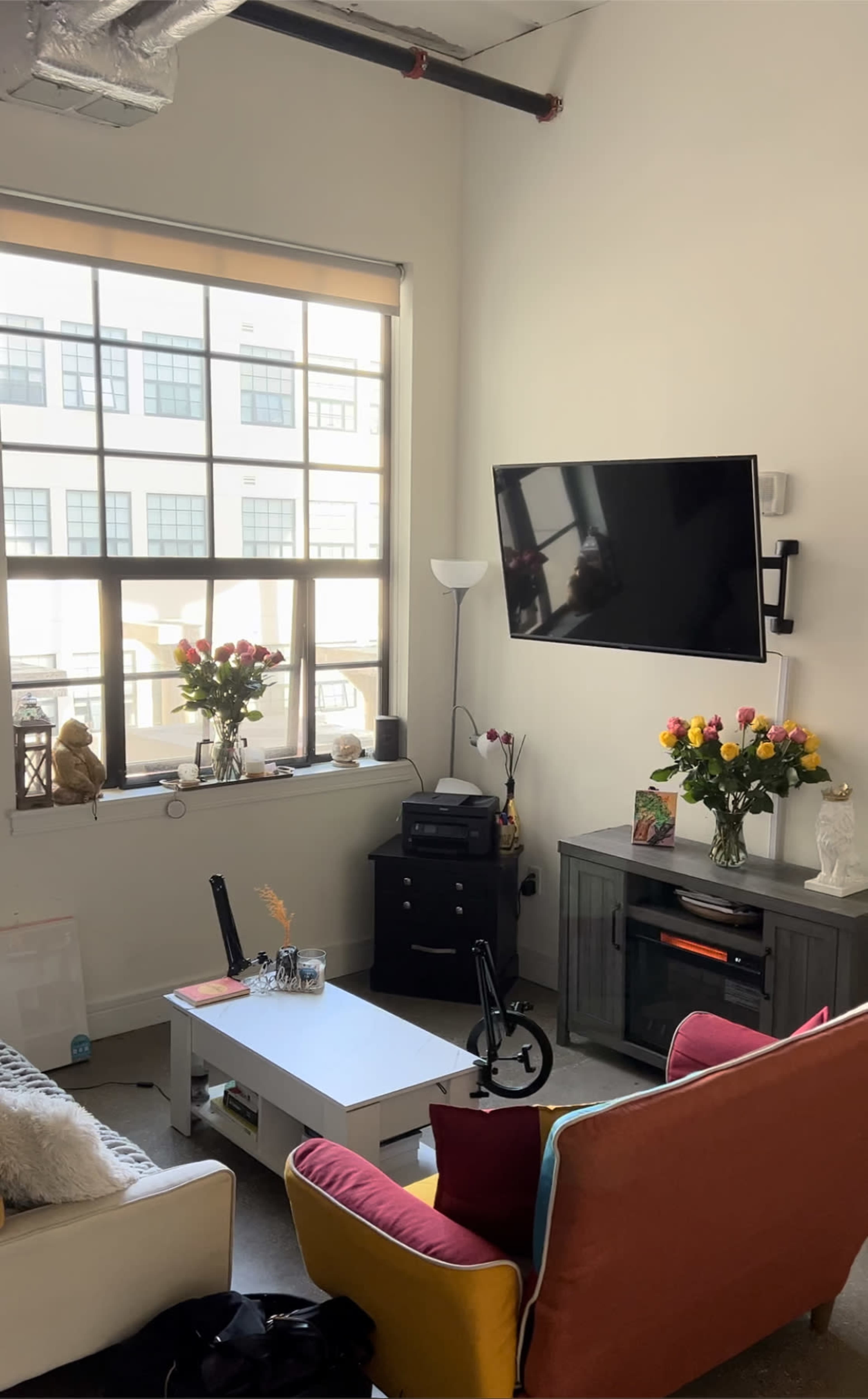 A modern living room with a large window, a wall-mounted TV, a coffee table, and colorful seating arrangements, along with flower arrangements on a side table.