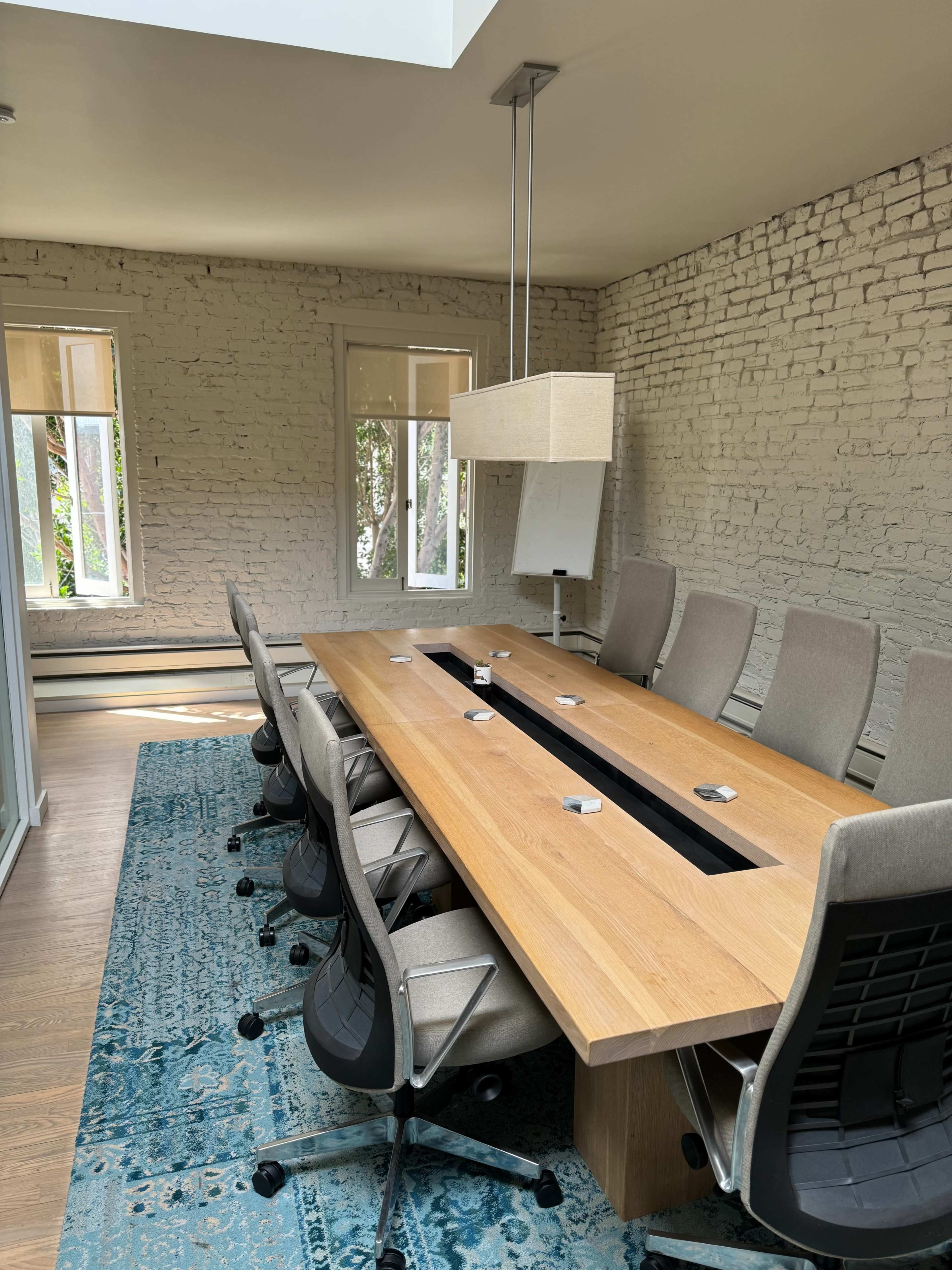 A modern conference room with a large wooden table, ergonomic chairs, and a light fixture overhead, all set against a backdrop of white-brick walls and large windows.