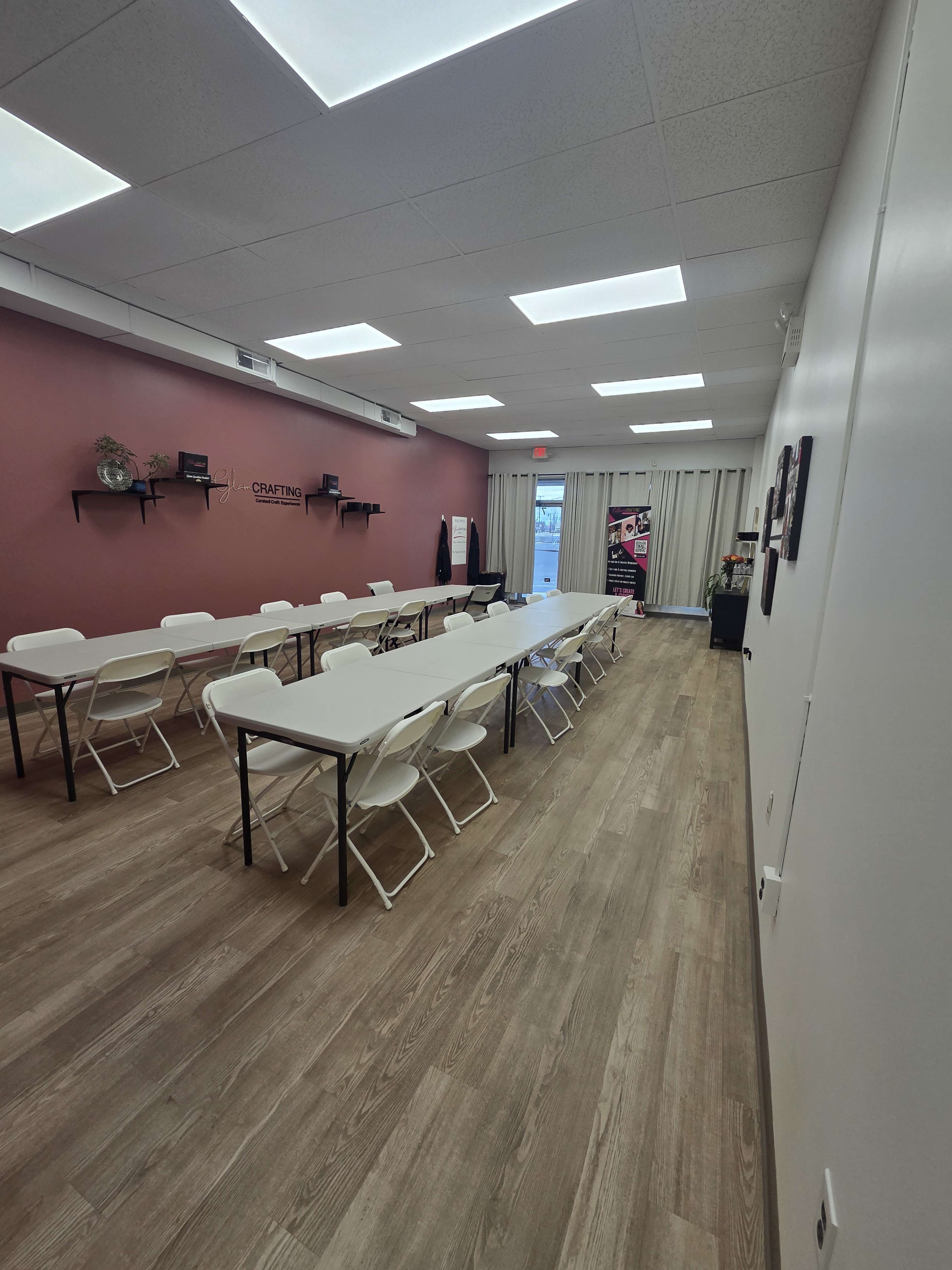 The image shows a spacious, empty room with multiple folding tables and chairs arranged in two rows, against a backdrop of a solid wall adorned with shelves and a light curtain covering a glass door.