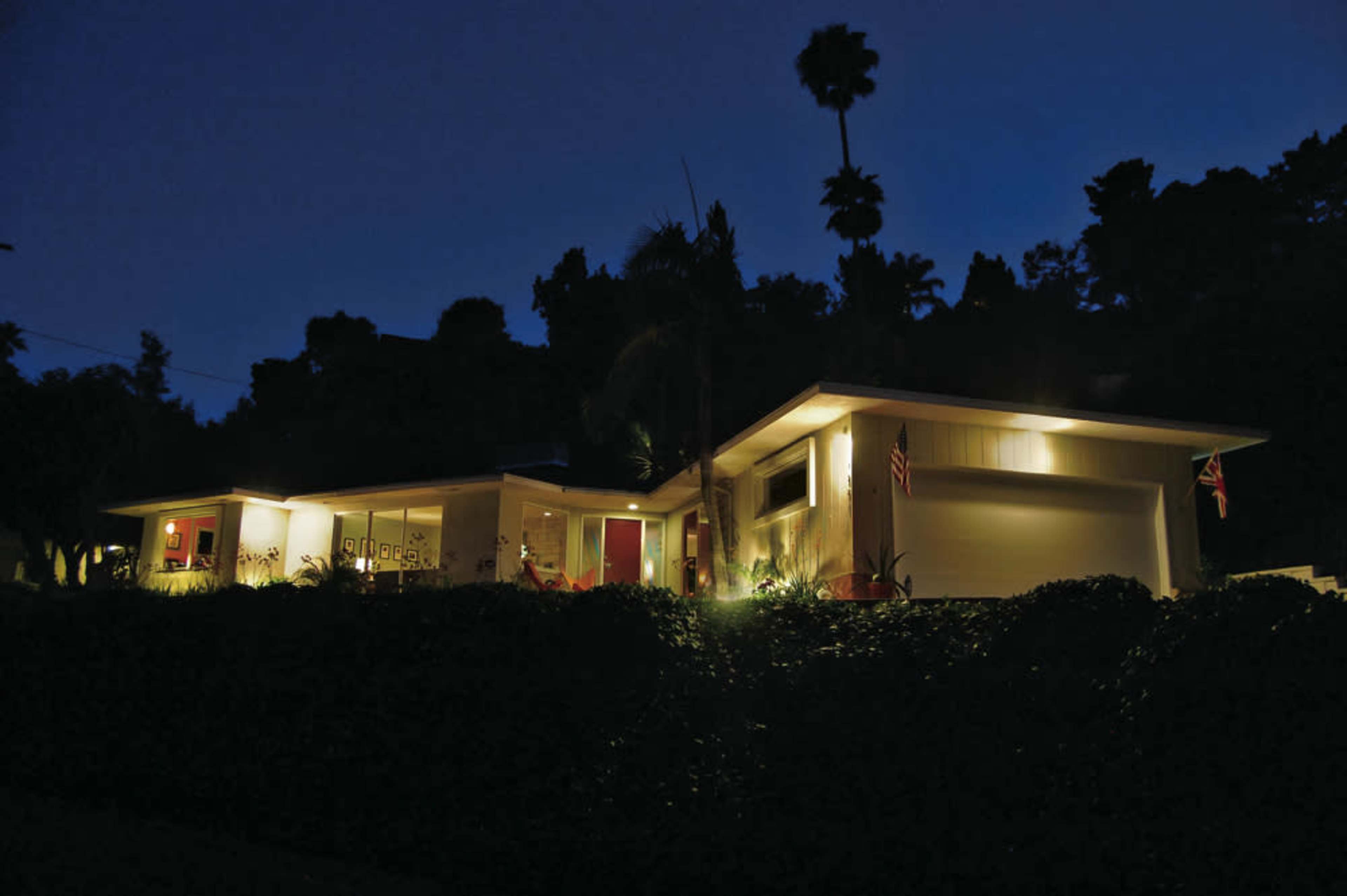 A modern house is illuminated at night, showcasing its architectural design and landscaped surroundings.