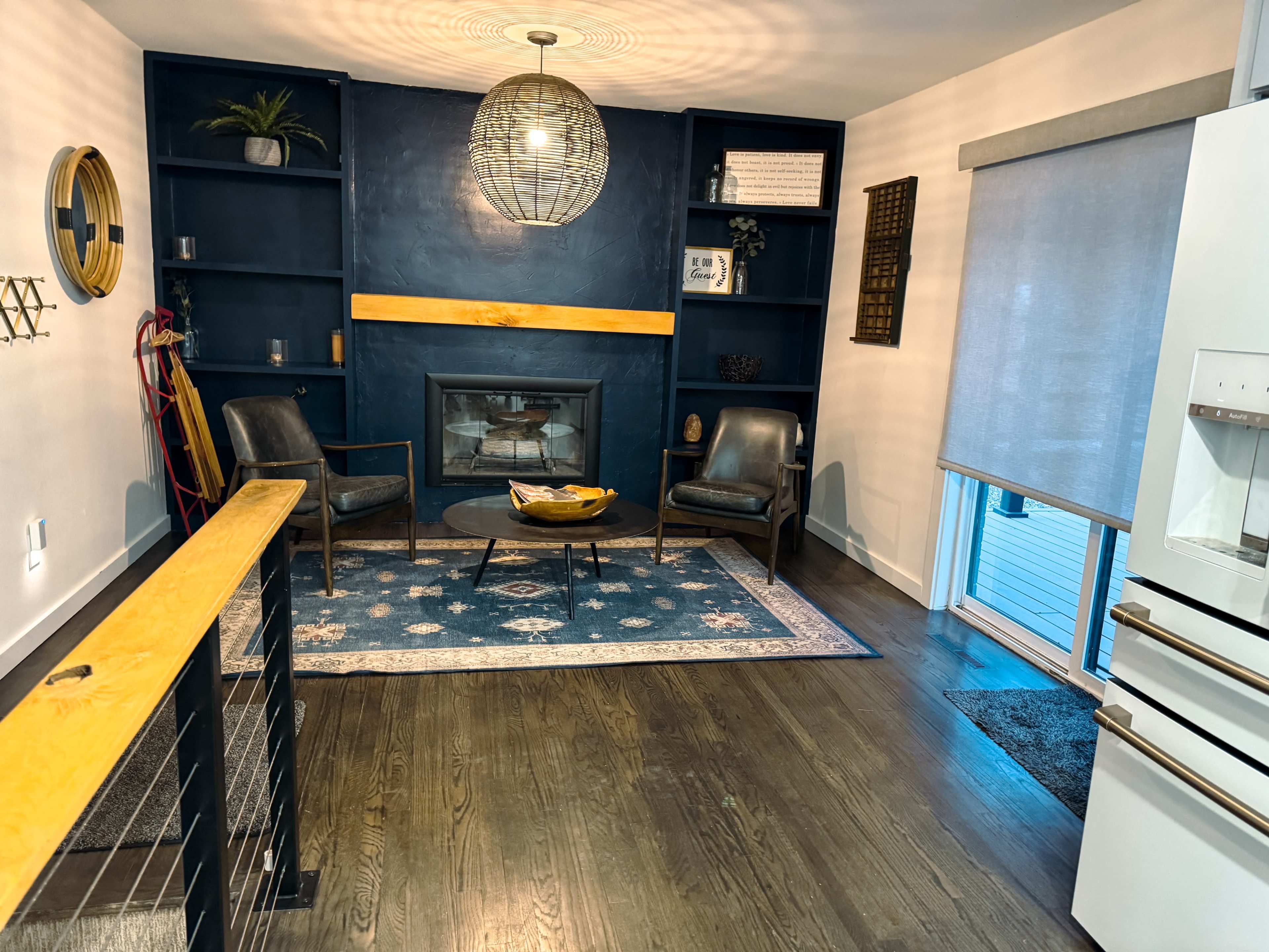The image shows a cozy living area with two chairs, a round coffee table, and a decorative bookshelf, centered around a fireplace.