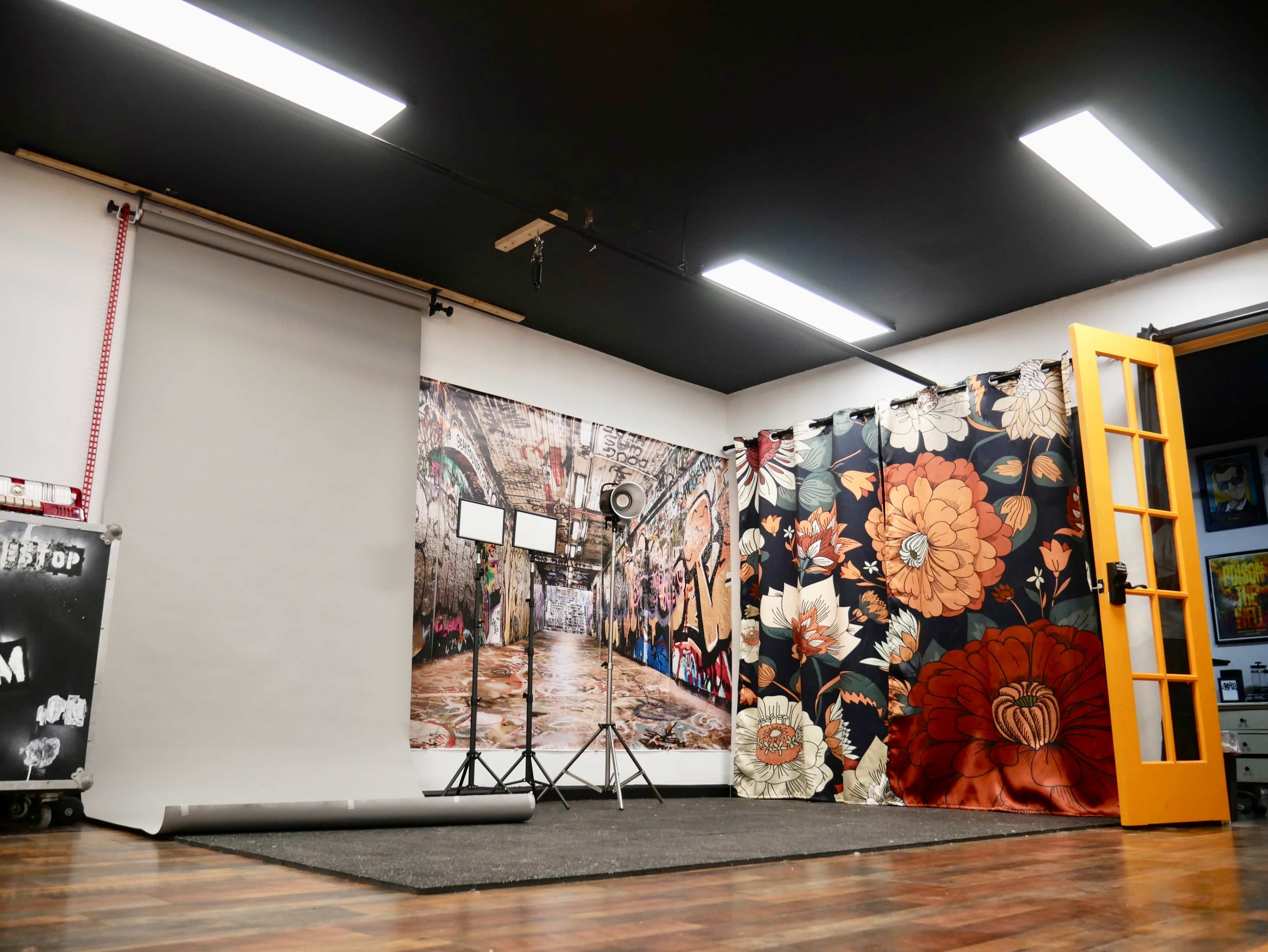 A photography studio features a backdrop of urban graffiti art alongside floral-patterned curtains, with studio lights on stands and a roll of backdrop paper on the floor.