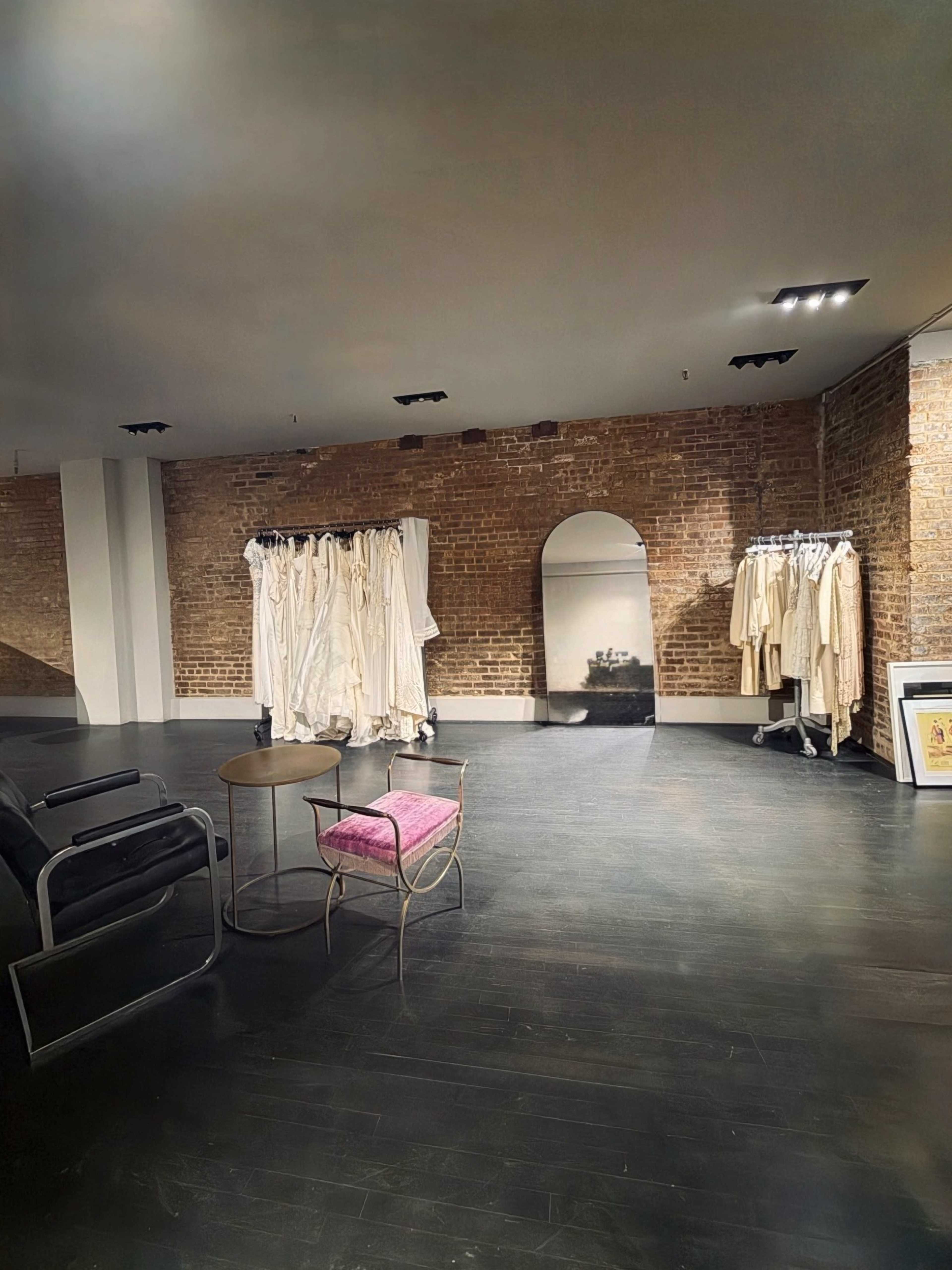 The image depicts a spacious retail interior featuring exposed brick walls, a clothing rack with white garments, a large mirror, and a black chair with a pink cushion.