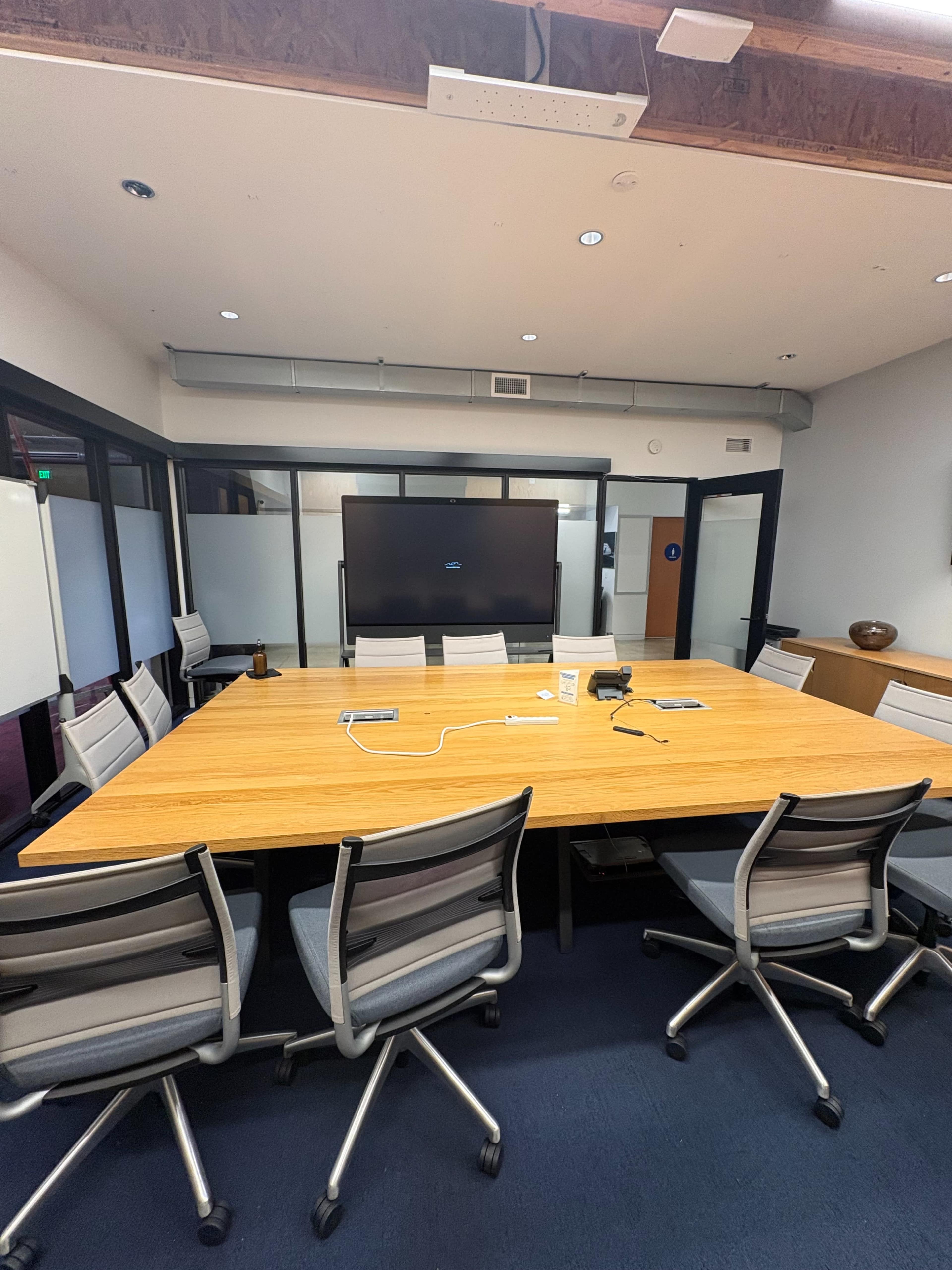 A large conference table surrounded by several chairs is set up in a modern meeting room, with a large screen displayed on one wall.