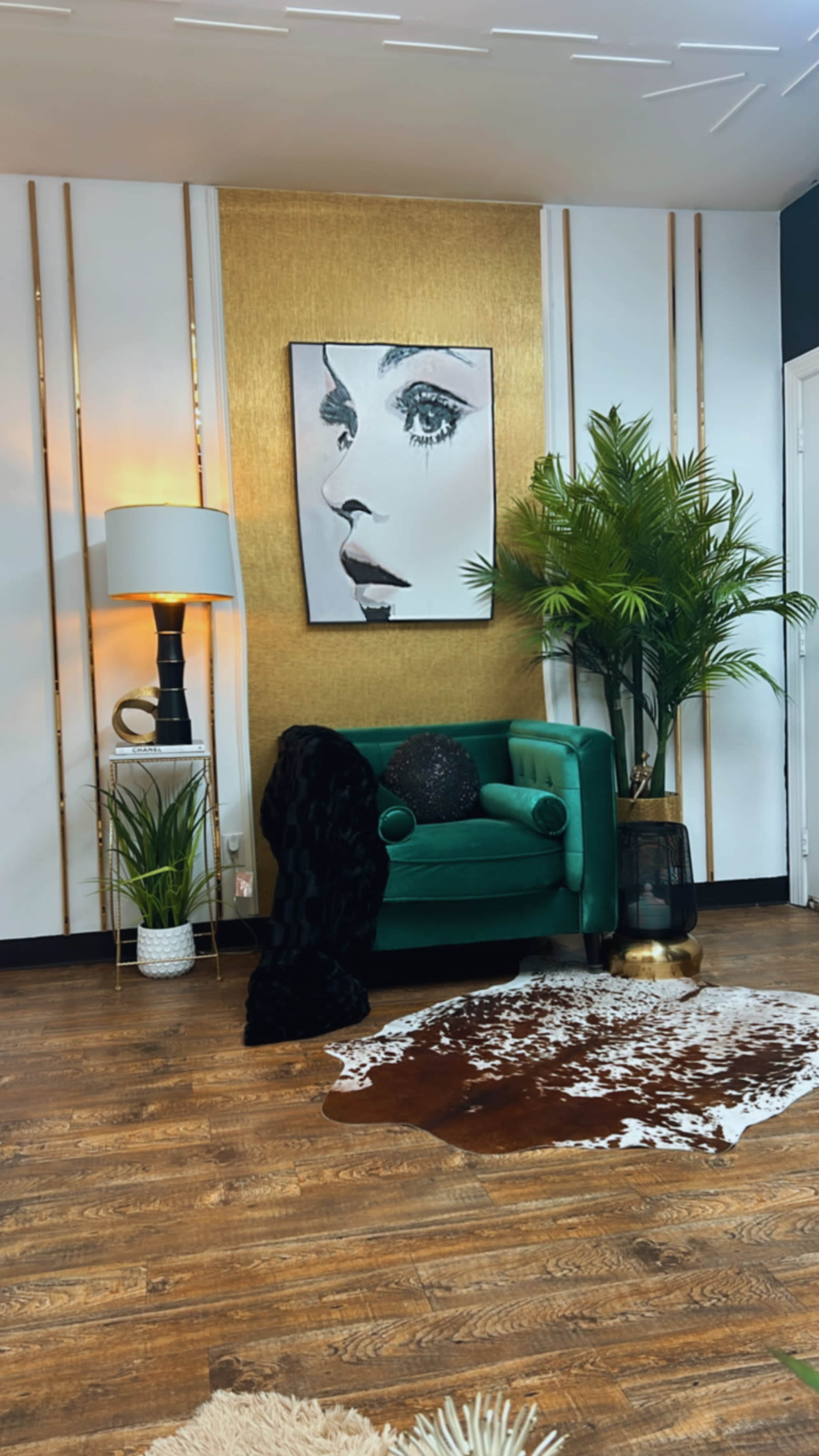 The image shows a stylish interior featuring a green velvet chair, a large portrait on the wall, a lamp, and decorative plants, with a cowhide rug on a wooden floor.