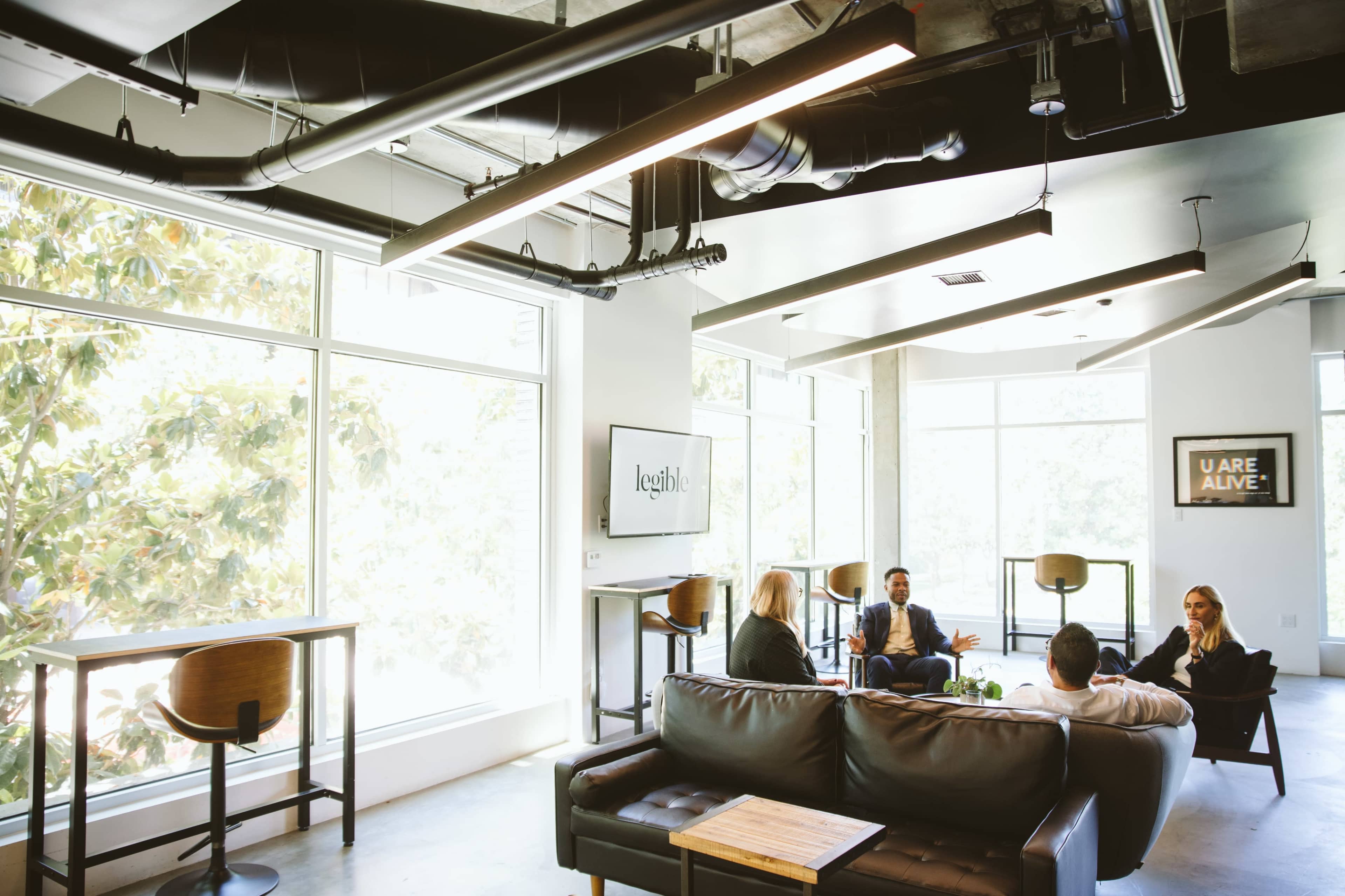 A group of four people engage in a discussion in a modern office lounge with large windows and industrial-style decor.