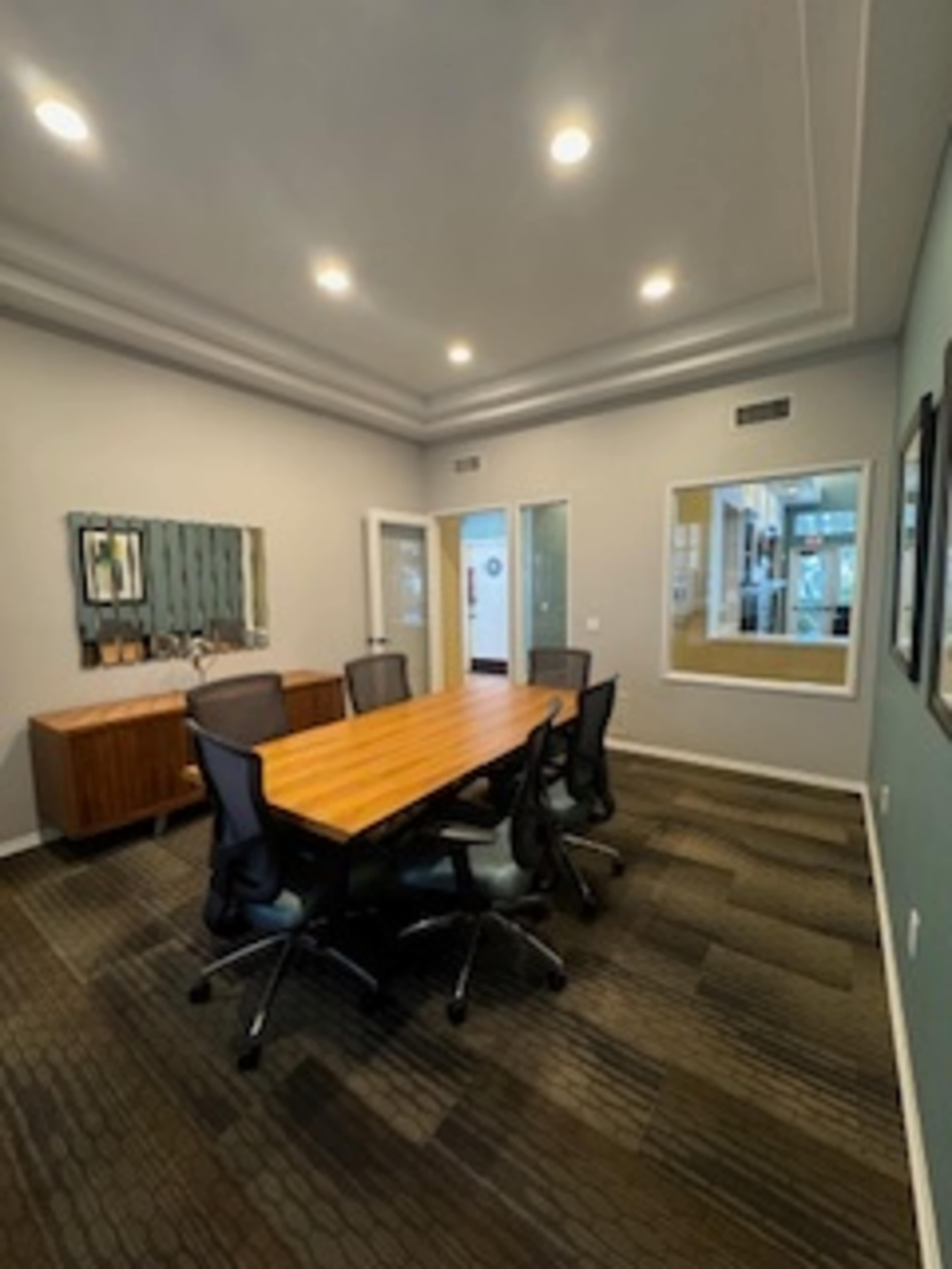 A modern conference room with a long wooden table surrounded by black chairs, and a large mirror on one wall.