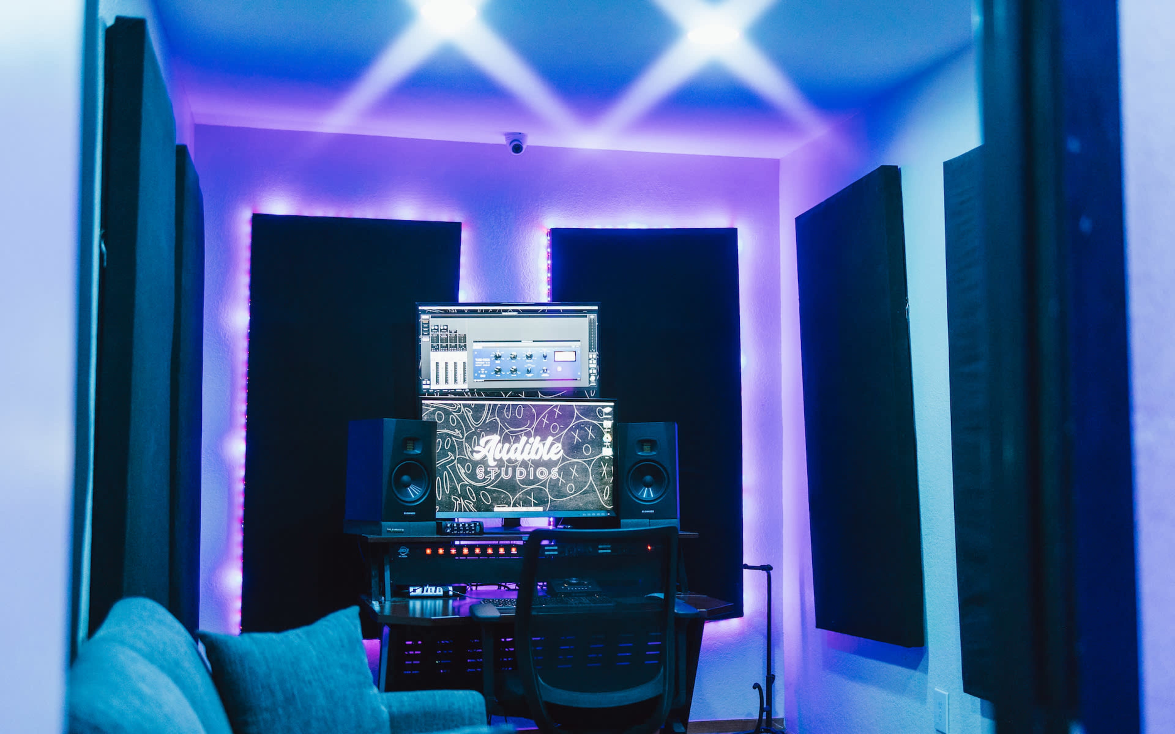 A music recording studio features dark soundproof panels and a computer screen with audio software, illuminated by purple LED lights.