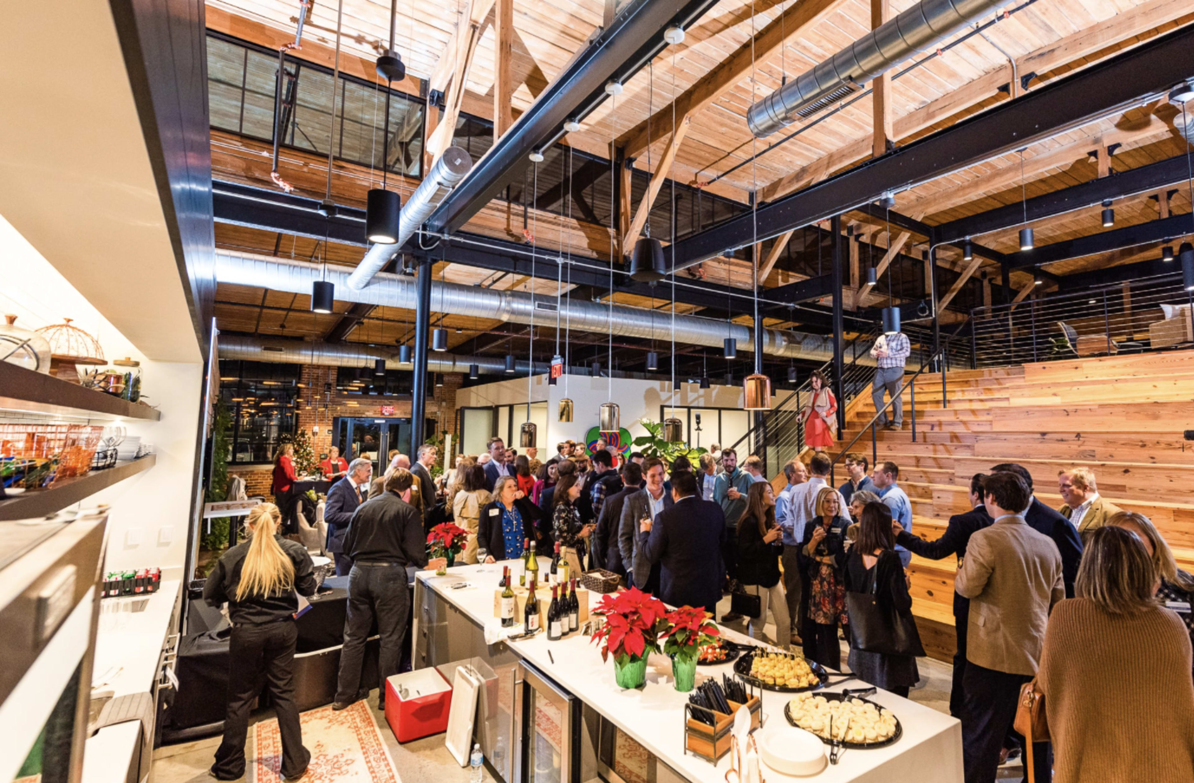 A group of people socializes at a spacious venue with exposed wooden beams and a bar area, surrounded by tables of food and drinks.