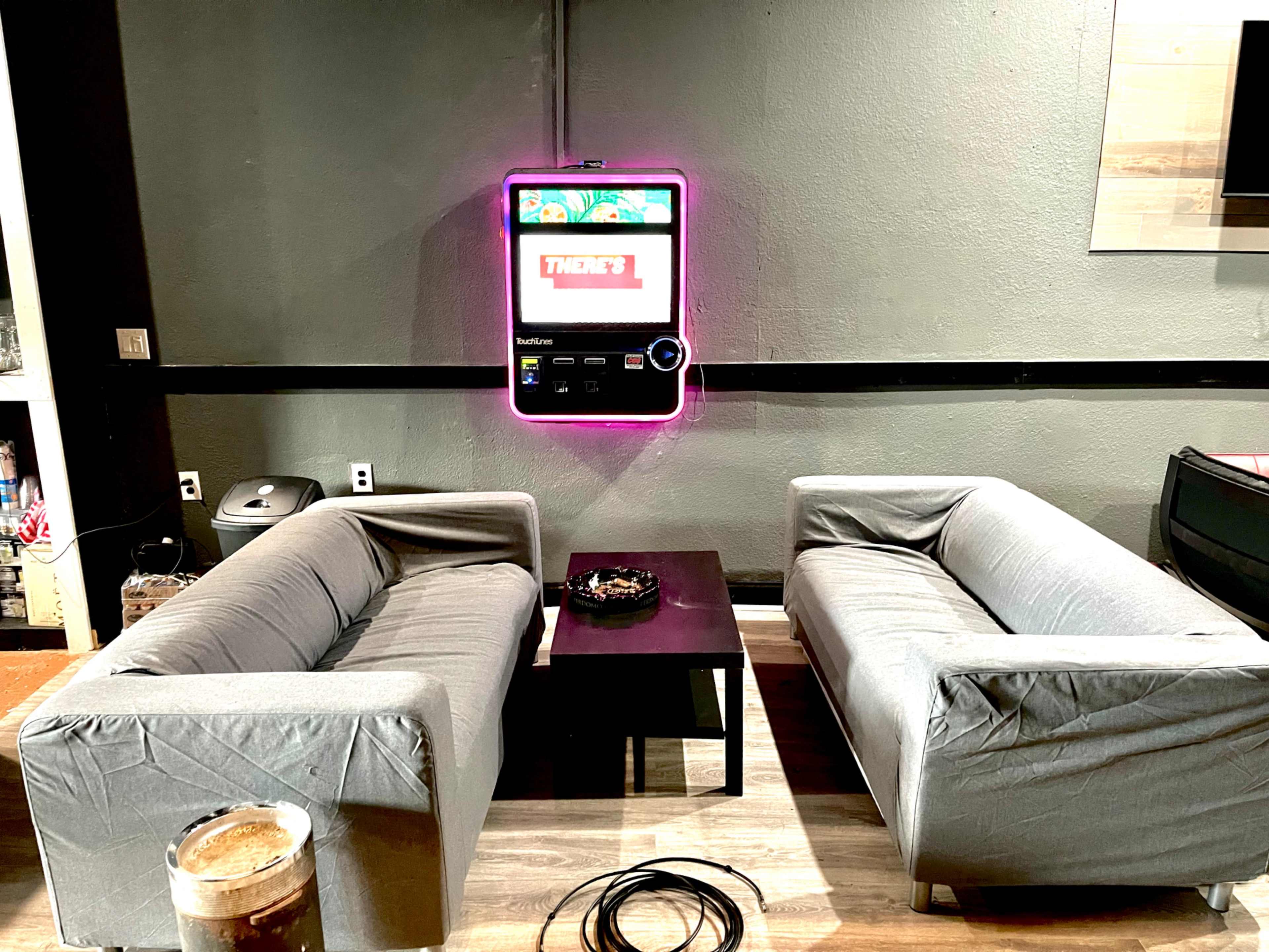 The image shows a minimalistic lounge area with two grey couches facing a small black coffee table and a neon-lit touchscreen kiosk on the wall above.