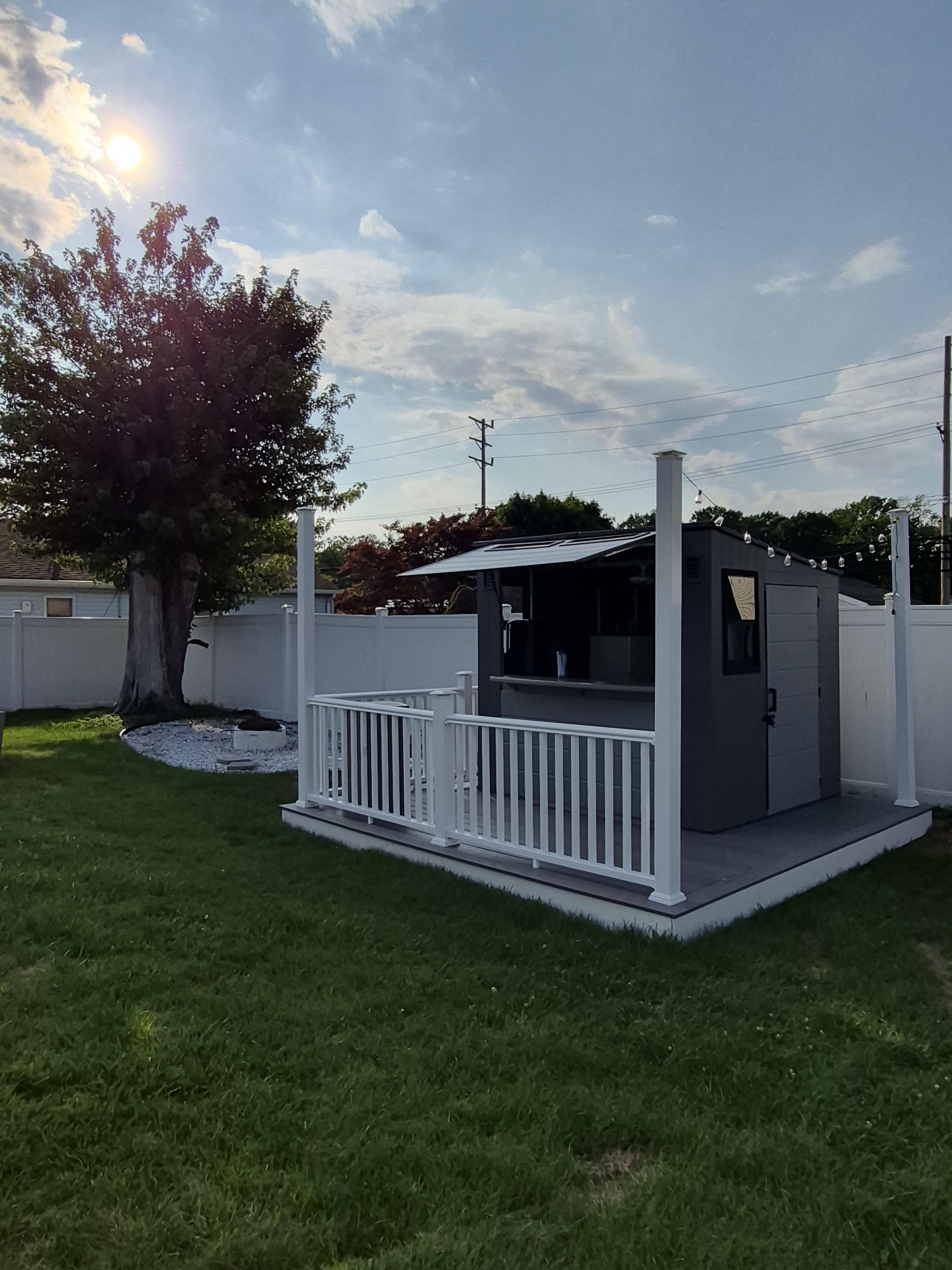 A small, gray outdoor shed with a porch is set in a grass yard, surrounded by a white fence and a large tree, with the sun shining in the background.