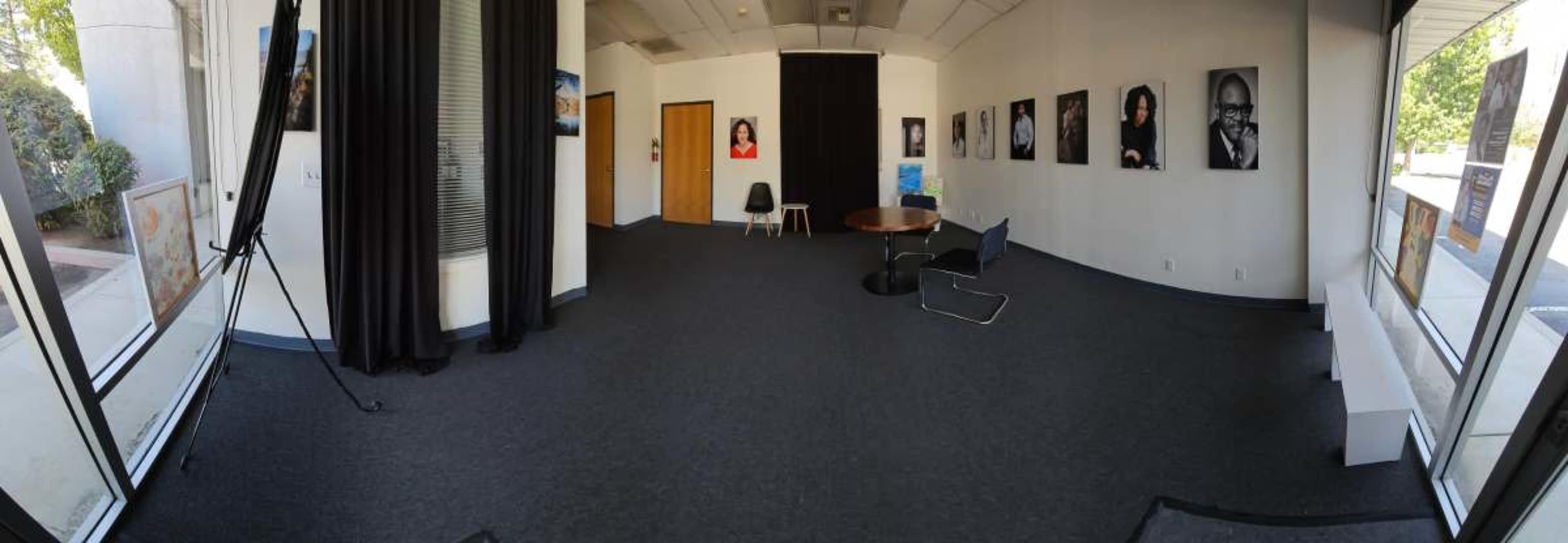 The image shows a spacious gallery with black curtains, a round table, and several framed photographs displayed on the walls.