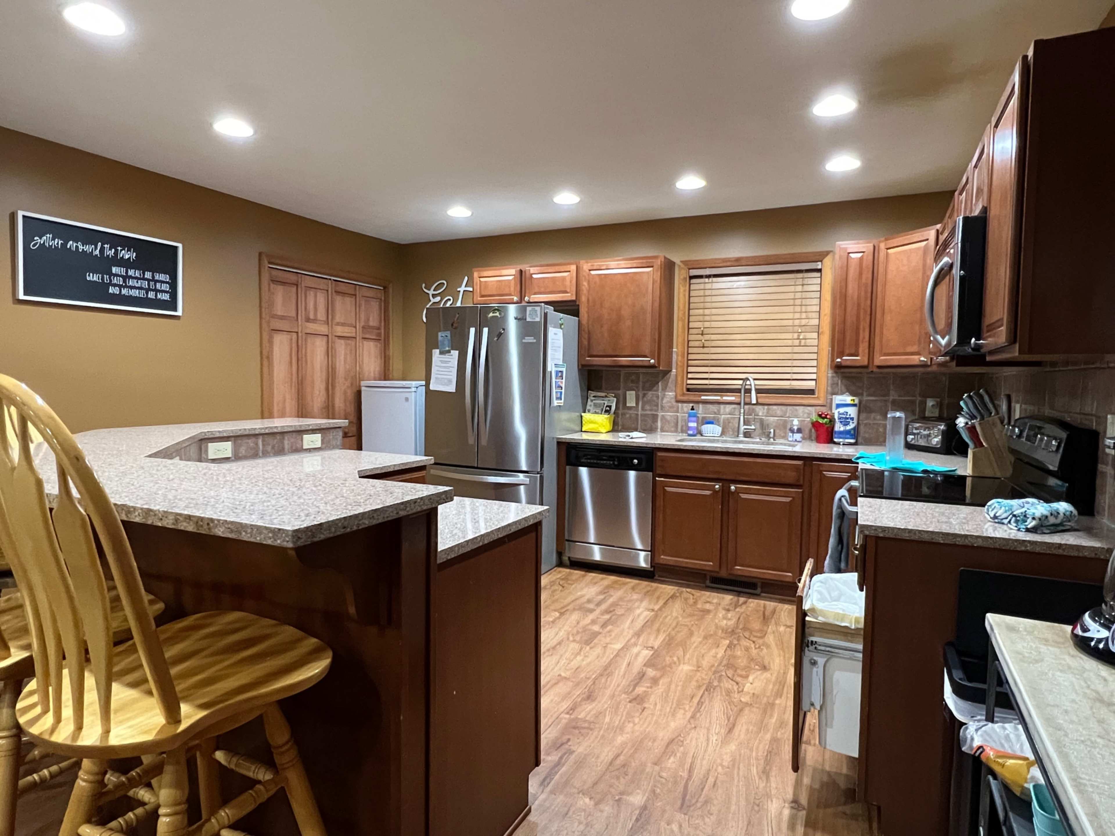 The image shows a kitchen with wooden cabinets, a stainless steel refrigerator, and a countertop with bar seating.
