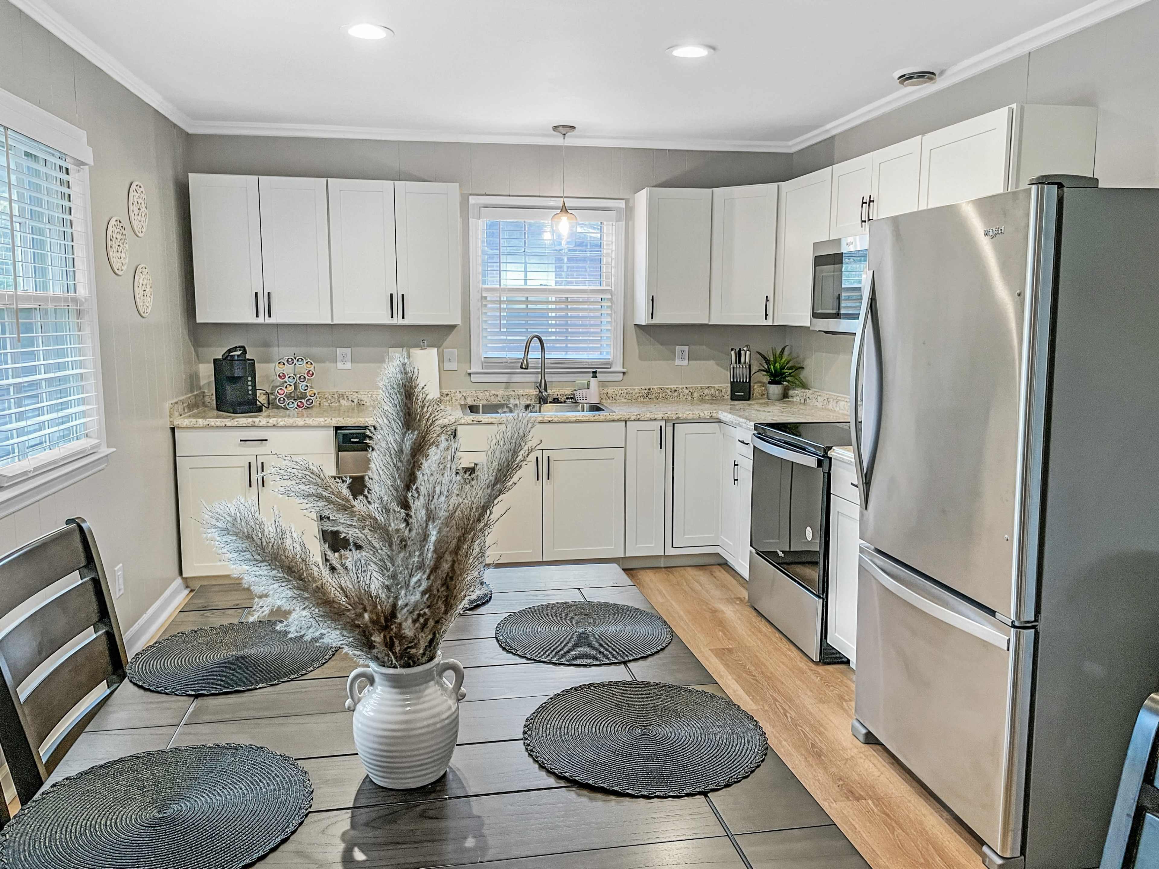 A modern kitchen features white cabinetry, stainless steel appliances, a kitchen island with a black table setting, and a vase of dried plants.
