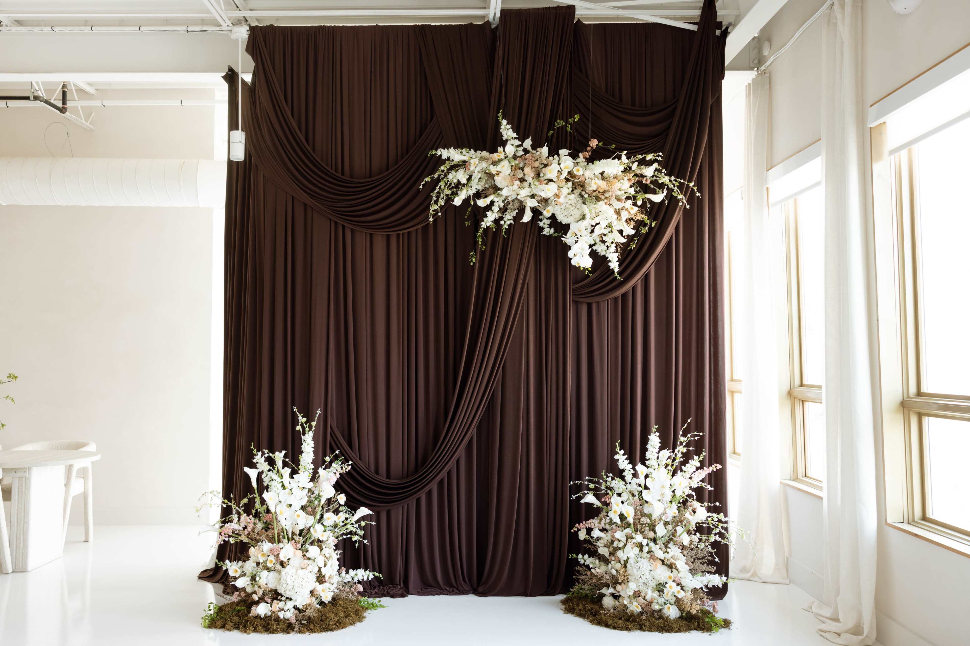 The image features a decorative backdrop with flowing brown drapery and an arrangement of white flowers on top, flanked by two floral arrangements on the ground.