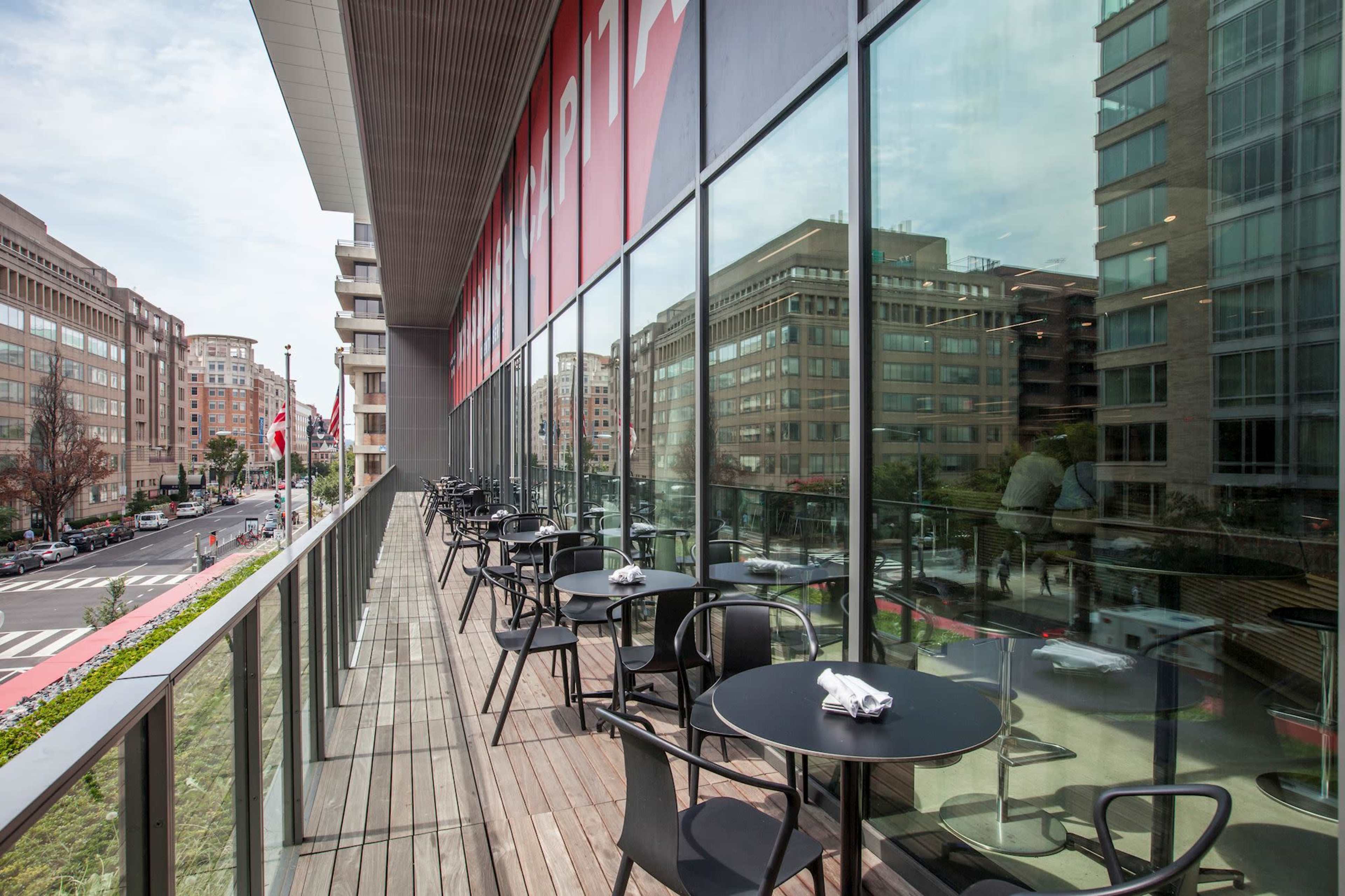 A modern outdoor dining area with black tables and chairs overlooks a city street lined with buildings.