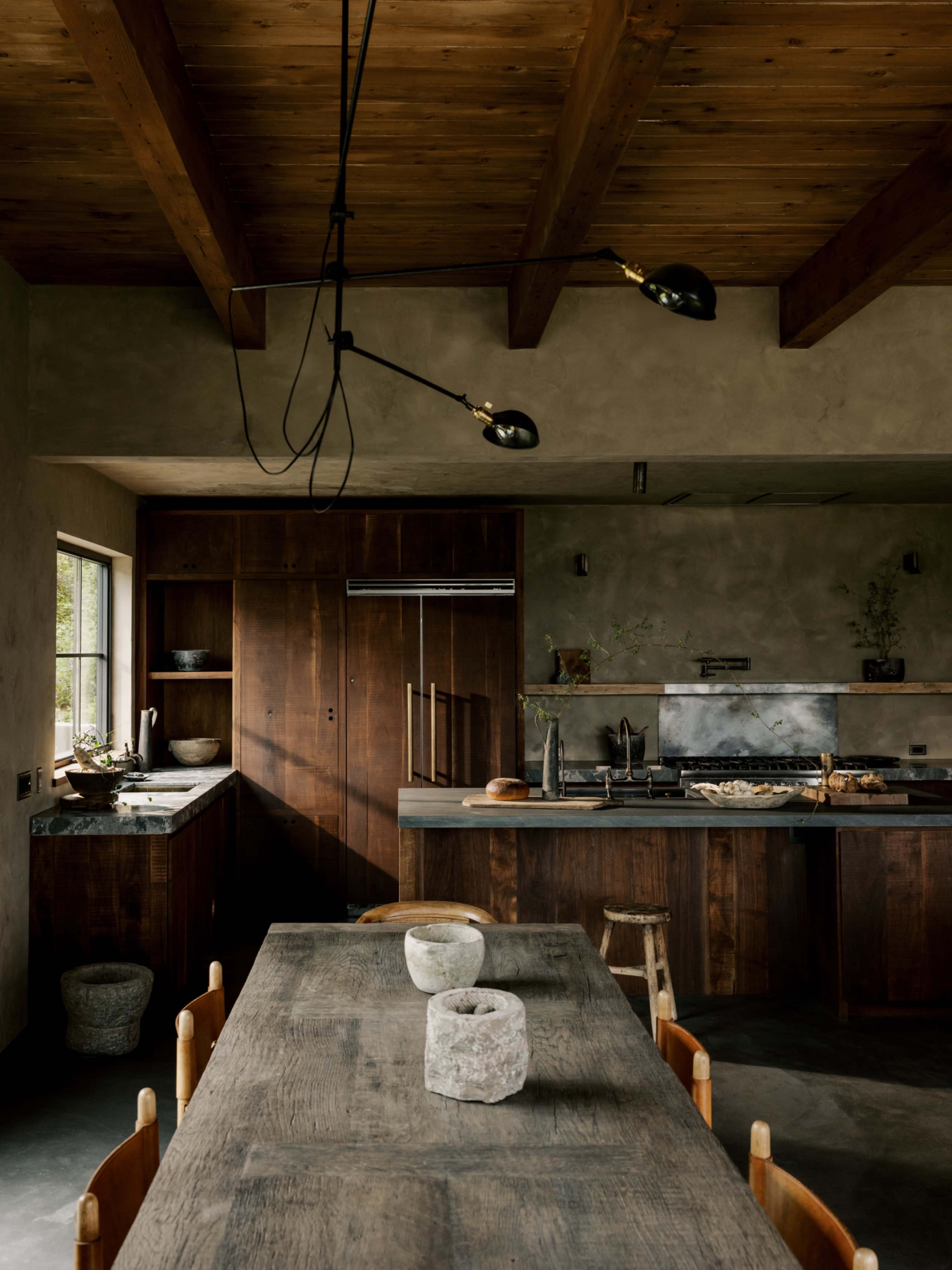The image shows a modern kitchen with wooden cabinetry, a large dining table, and industrial lighting fixtures.